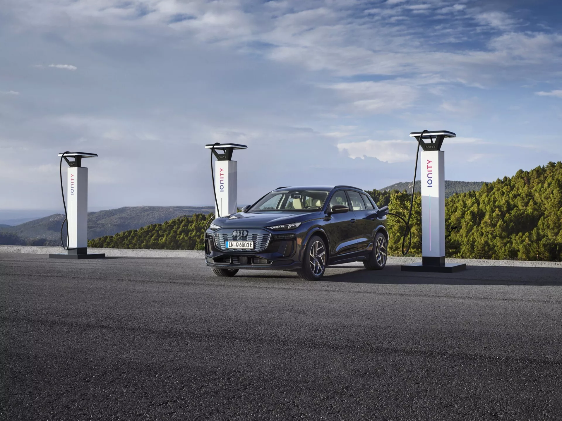 SUV Audi Q6 e-tron 2024 v hluboké metalické modři u nabíjecí stanice pro elektromobily se třemi nabíječkami. Obrázek zachycuje vozidlo v úhlu tří čtvrtin, ukazující jeho přední levou stranu, zvýrazňující mřížku, světlomety, kola a kapotu. Stylové a moderní elektrické vozidlo vykazuje sportovní a všestranný design s elegantními agresivními světlomety a litými koly, zdůrazňující pokročilou a udržitelnou automobilovou technologii.