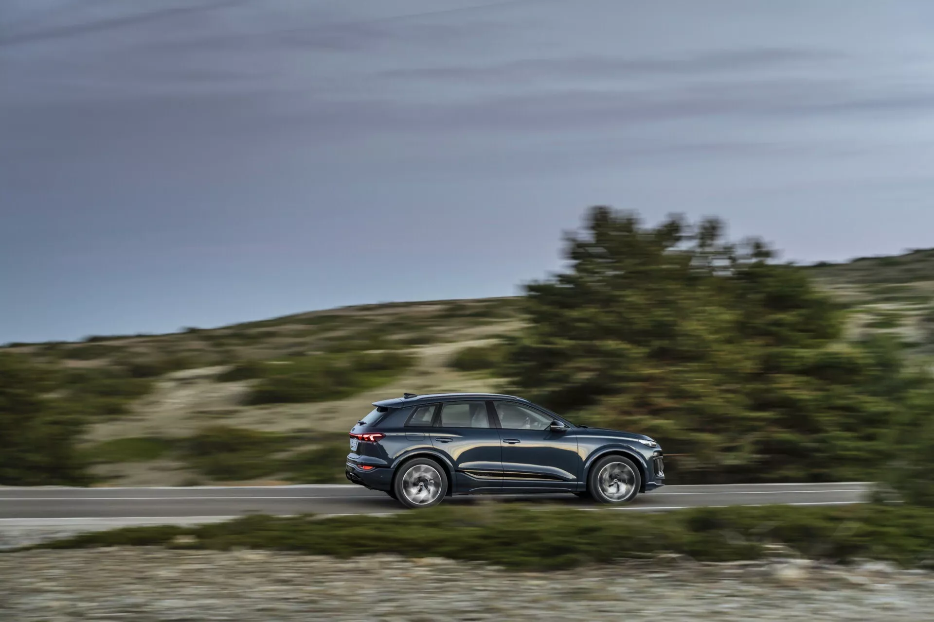Audi Q6 e-tron 2024 dynamický boční pohled v pohybu s rozmazaným pozadím, střední paralelní úhel zobrazující profil, designové linie, okna, pět dveří, boční zrcátka, hliníkové ráfky, denní světlo, venkovské pozadí.