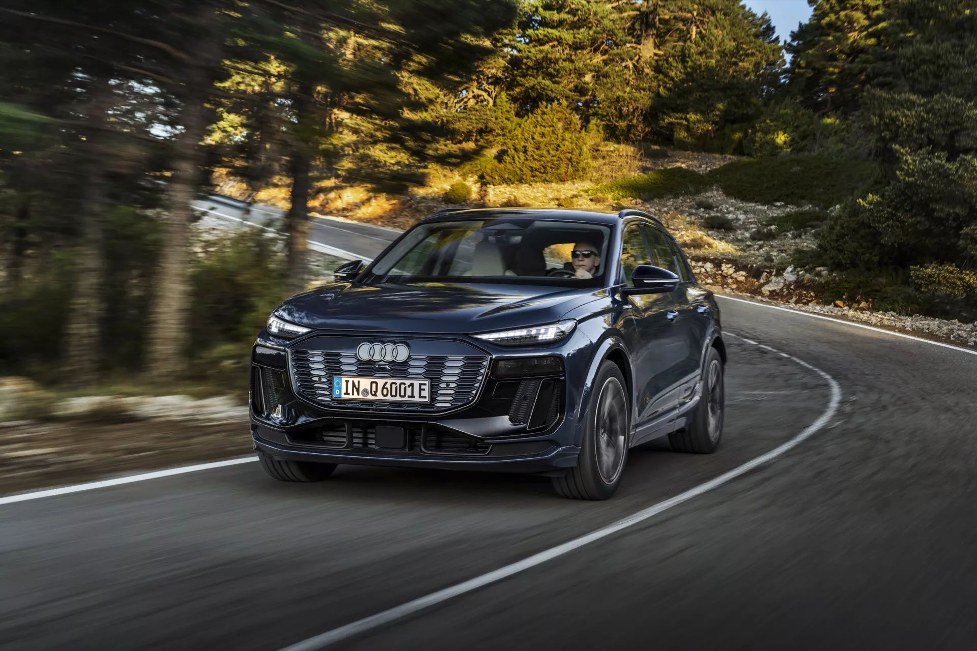 Obrázek moderního SUV Audi Q6 e-tron 2024 v pohybu, zachycený z dynamického tříčtvrtečního předního úhlu, zvýrazňující design a výkon. Vozidlo, identifikované podle charakteristického loga se čtyřmi kruhy, má emblémová LED světla, agresivní přední nárazník a tvarované linie kapoty. Obrázek, pořízený z nižšího úhlu, zvýrazňuje přítomnost vozu na zakřivené silnici, přičemž jeho slitá kola jsou mírně rozmazaná, což naznačuje pohyb. Nový model naznačuje pokročilé technologie jako adaptivní tempomat a elektrický pohon, označené odznakem 'e-tron'. Profesionální fotografování, které představuje luxus, design a technologii v konfiguraci s levostranným řízením.