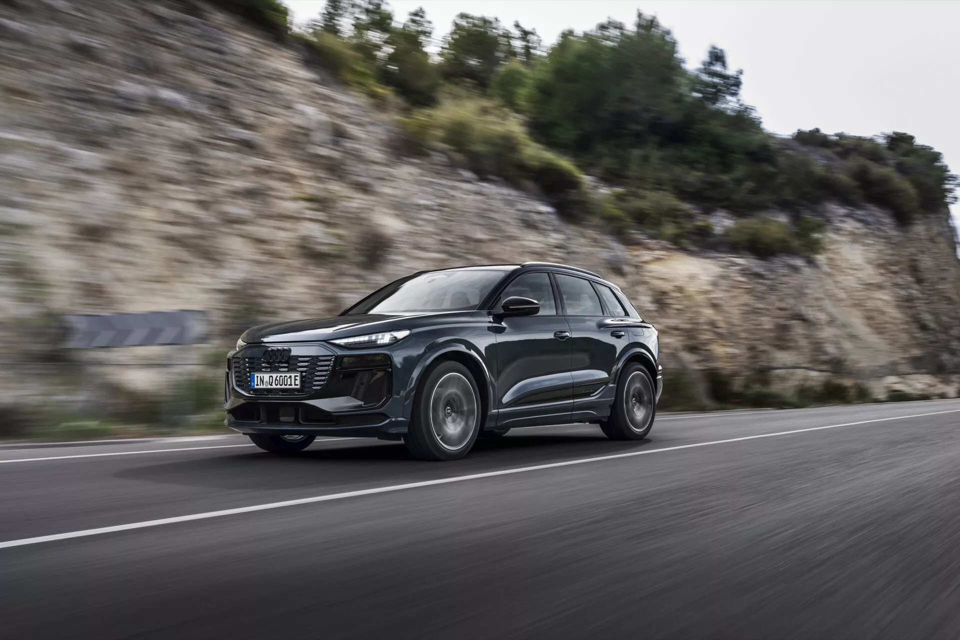Dynamický záběr Audi Q6 e-tron 2024 v pohybu z nízkého úhlu, čelně zepředu, zobrazující jeho elegantní aerodynamický design SUV, moderní LED světlomety a hliníková kola s rozmazáním pohybu, naznačující výkonové schopnosti na asfaltové cestě.