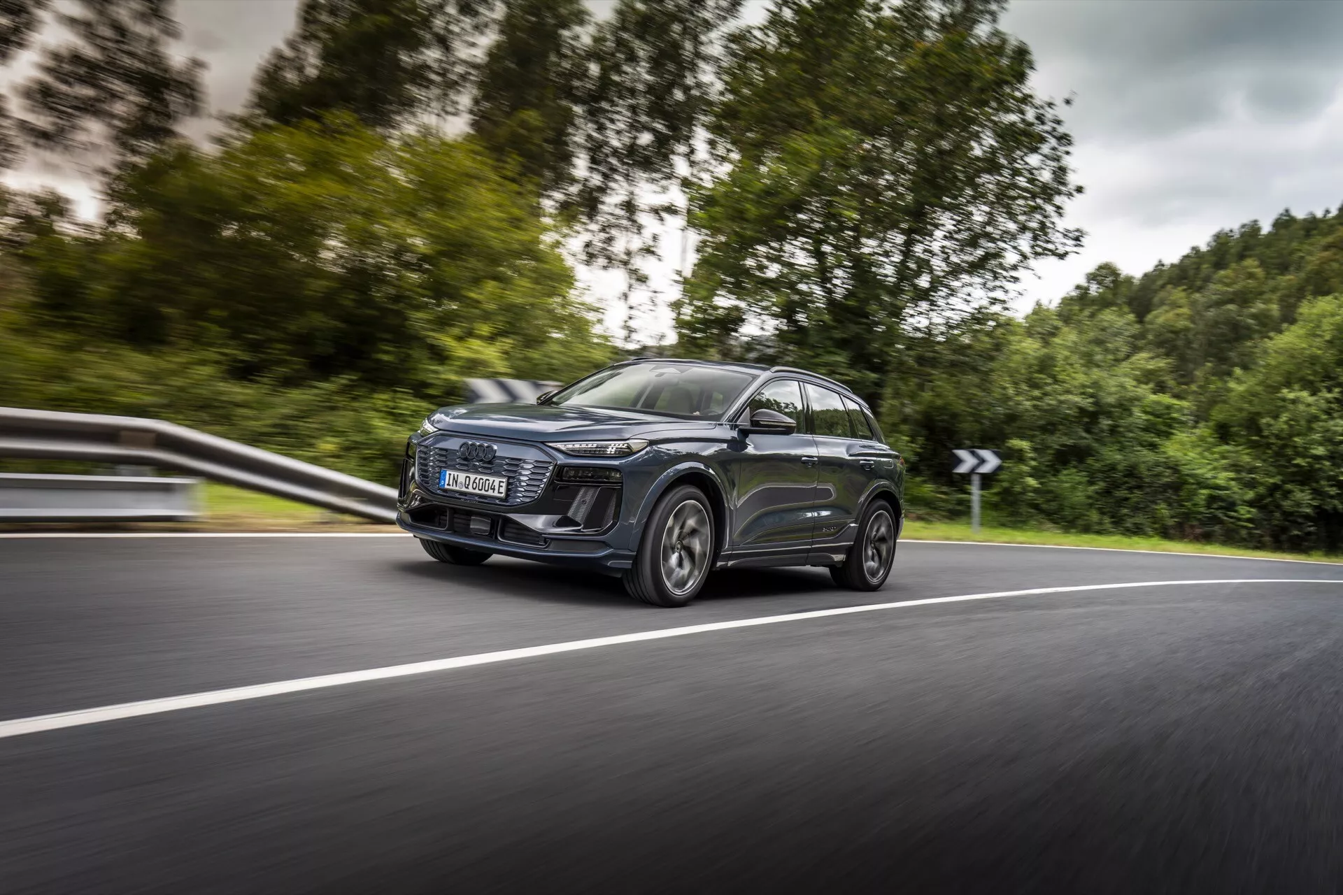 Obrázek zobrazuje moderní Audi Q6 e-tron 2024 SUV v pohybu, zachycený z úhlu zepředu zleva, soustředící se na jeho levou stranu. Dynamický snímek zdůrazňuje výrazné přední levé světlomet s charakteristickým LED denním svícením, velkou moderní mřížku, elegantní aerodynamická zrcátka s integrovanými směrovými světly a velká litá kola s nízkoprofilovými pneumatikami. Auto má panoramatické střešní okno a tónovaná skla, s reflexním metalickým lakem karoserie. Nízký úhel pohledu a mírné rozmazání pozadí zdůrazňují rychlost a pohyb auta na zakřivené dvouproudové silnici.