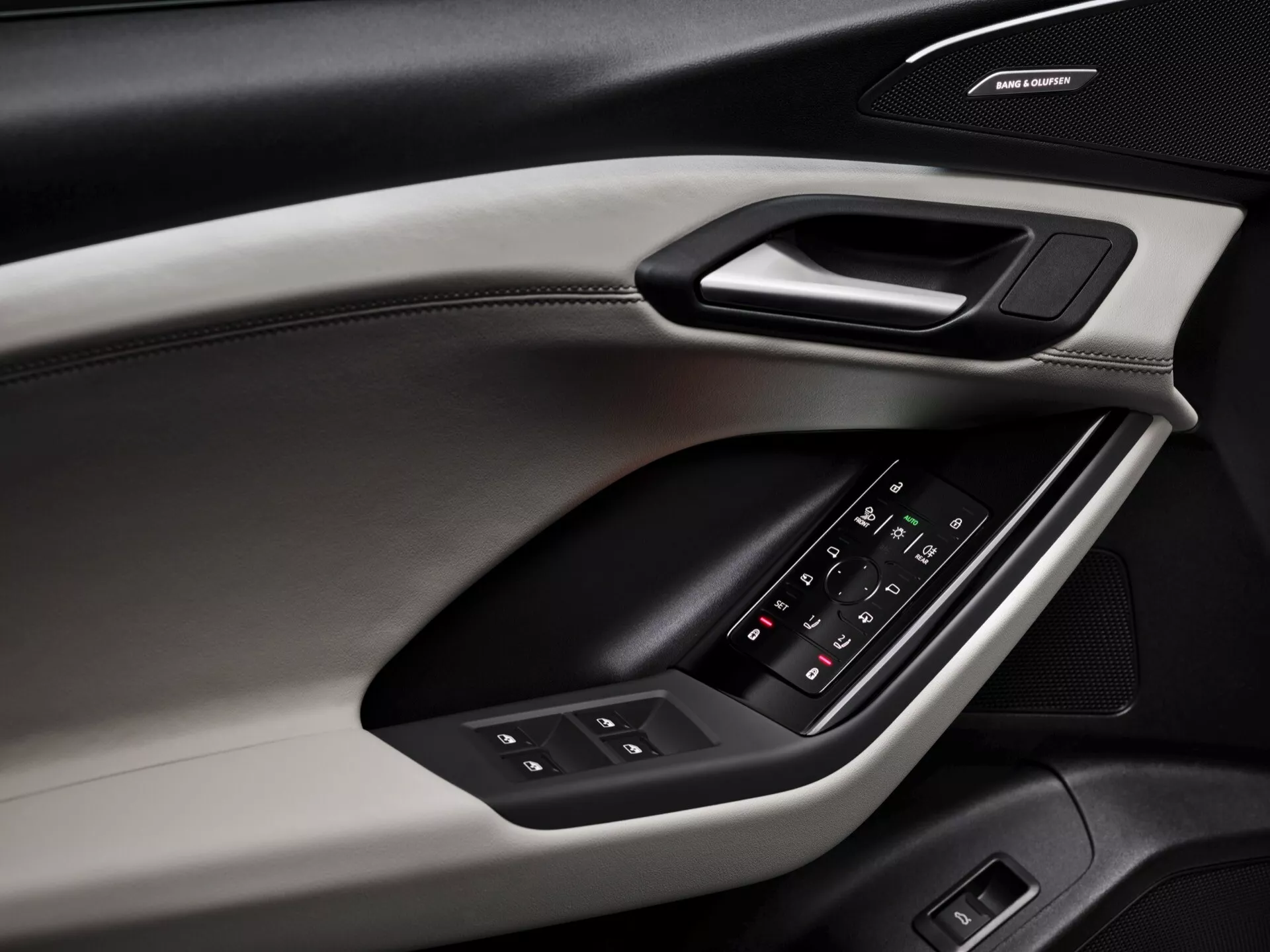Detailní pohled na vnitřní dveřní panel Audi Q6 e-tron 2024, zdůrazňující designové prvky a vybavení. Panel dveří, snímaný z úhlopříčného úhlu, odhaluje různé materiály a textury, včetně měkké horní části s viditelným prošitím, tvrdého obložení obsahujícího ergonomickou rukojeť a polstrovanou opěrku s podsvíceným ovládacím panelem pro okna a nastavení zpětných zrcátek. Pod opěrkou je značkový reproduktor, naznačující prémiový audio systém. Barevné schéma zahrnuje dvoutónový design, který zvyšuje estetické a funkční kvality.