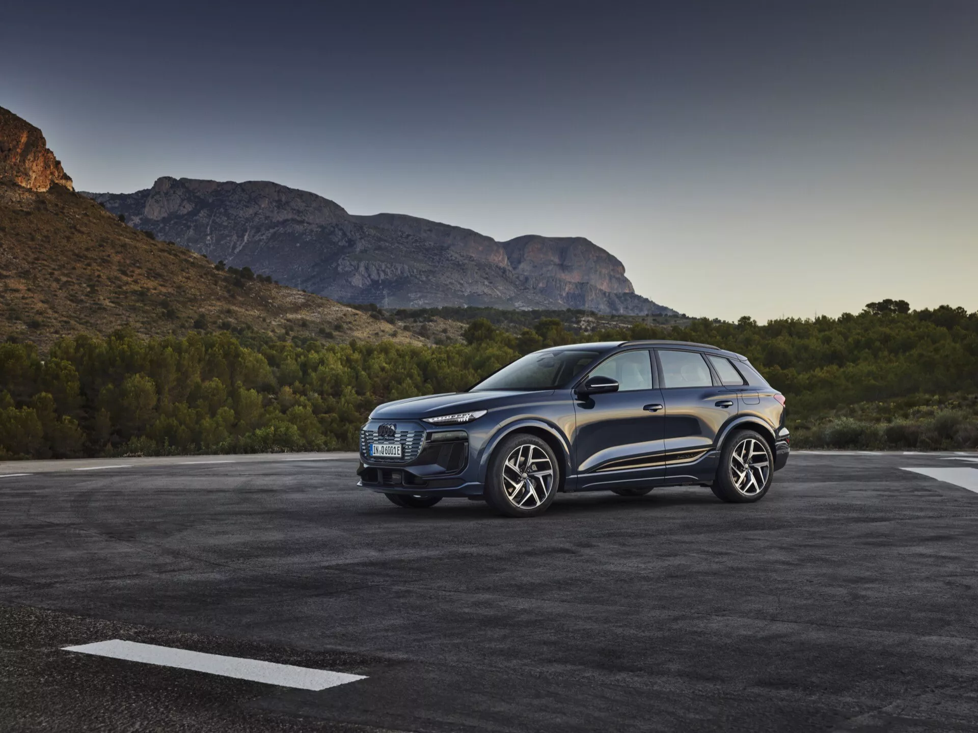Tříčtvrteční pohled zepředu na modrý SUV Audi Q6 e-tron 2024 zaparkovaný venku, který ukazuje výraznou masku, elegantní světlomety, tvarovanou kapotu, velká kola z lehké slitiny, boční zrcátka a dveře na straně řidiče na pozadí malebné horské krajiny za ranního nebo večerního světla.