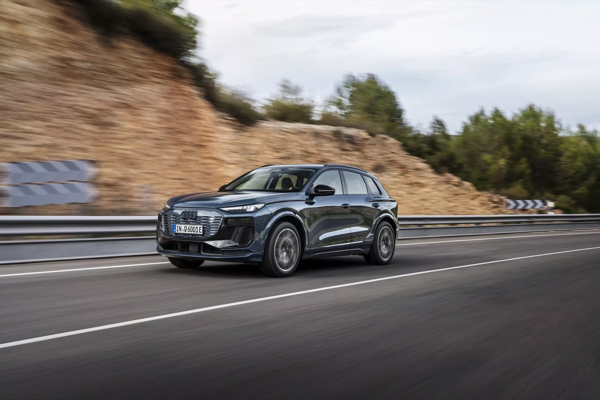 Obrázek Audi Q6 e-tron 2024 v pohybu, pořízený technikou panningu, která zdůrazňuje elegantní profil a moderní design SUV. Auto, rámované z bočního úhlu, se řítí po víceproudé dálnici s rozmazanými koly a pozadím, což podtrhuje jeho dynamické schopnosti. Tmavě kovové vozidlo má velkou přední masku, výrazné LED světlomety, sportovní hliníková kola a reflexní povrchy. Zatažená obloha a mokrá vozovka vytvářejí dramatickou scénu, zdůrazňující luxusní a výkonné funkce vozu.