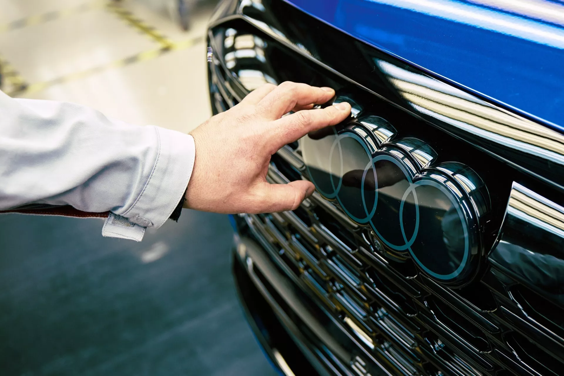 Detailní pohled na přední mřížku Audi Q8 e-tron 2023 se zaměřením na znak a ruku dosahující k němu, což zdůrazňuje potenciální technologické funkce a prémiový vnější design