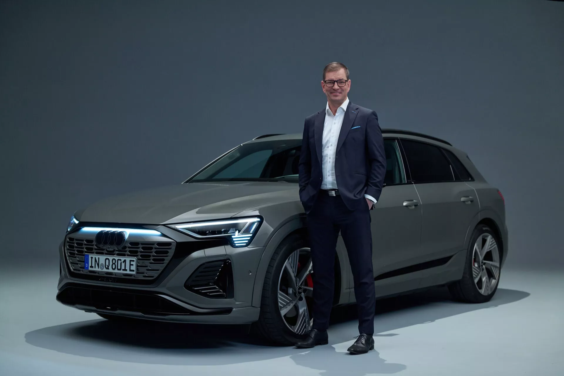 Audi Q8 e-tron 2023 v prostředí studia, prezentující přední mřížku, LED světlomety, vícepaprsková slitinová kola a elegantní SUV karoserii s mužem v obchodním oblečení pro měřítko