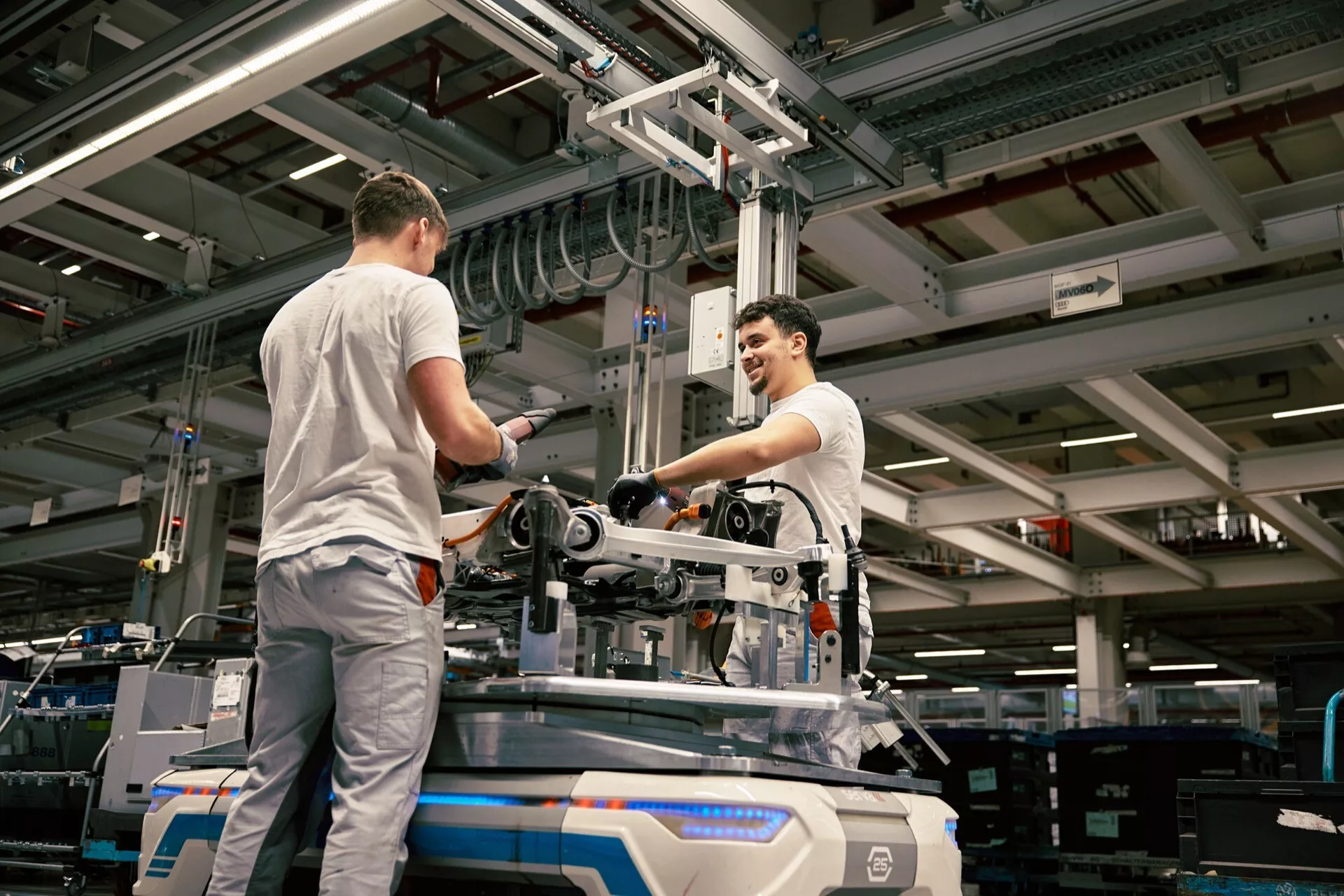 Průmyslové prostředí se dvěma pracovníky montujícími nebo údržbou mechanického vybavení na podlaze továrny, pod umělým osvětlením, s vyvýšeným pohledem zdůrazňujícím výrobní prostředí