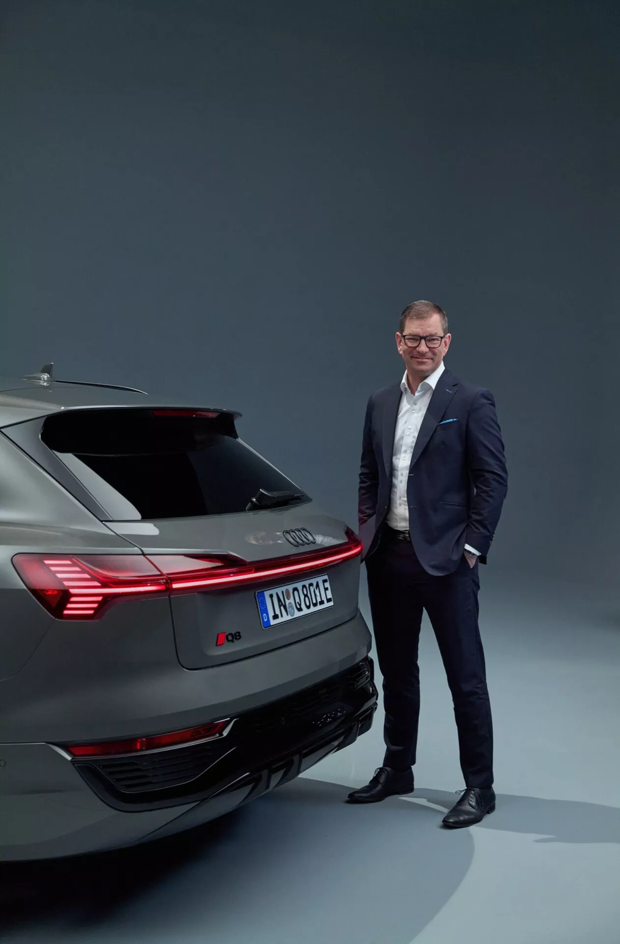 Audi Q8 e-tron z roku 2023 ukazující zadní design včetně zadních světel, nárazníku a systému výfuku v ateliérovém nastavení, s profesionálně oblečeným mužem stojícím vedle, což zdůrazňuje moderní automobilovou technologii a luxus.