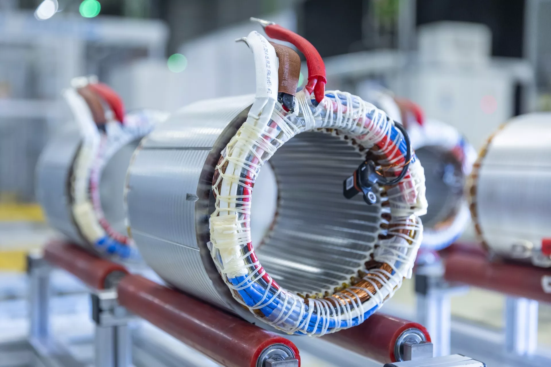 Fotografie ve vysokém rozlišení statoru elektrického motoru umístěného na červeném nosiči, zobrazující cívky drátu navinuté kolem laminovaných kovových jader typických pro elektrické stroje pro generování magnetických polí