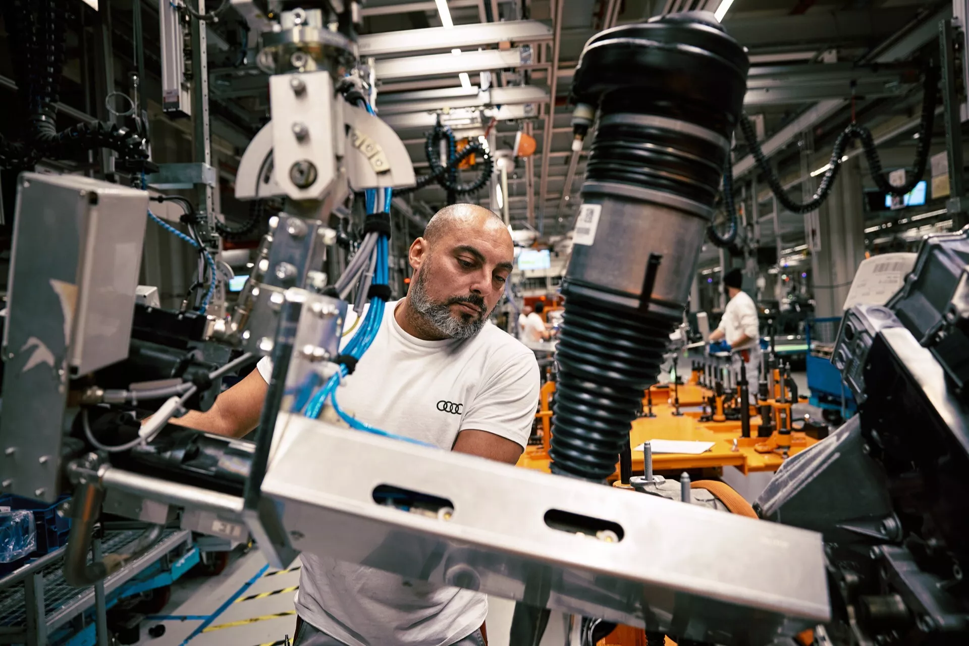 Pracovník kontroluje nebo montuje komponentu pérování automobilu, možná tlumič, na výrobní lince Audi Q8 e-tron 2023 se zamazaným pozadím strojů