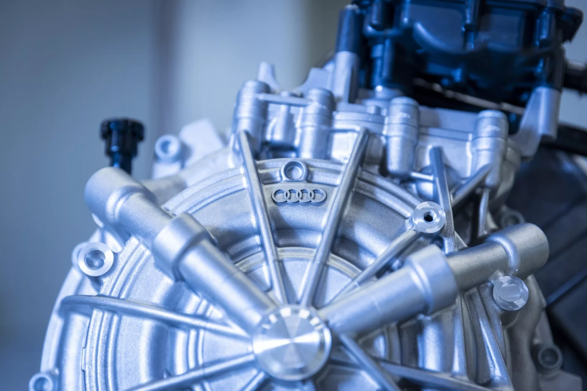 Detailní pohled na komponent motoru Audi Q8 e-tron 2023, zobrazující kruhový střed s vyzařujícími prvky a logo Audi, předvádějící pokročilé automobilové inženýrství a precizní výrobu.