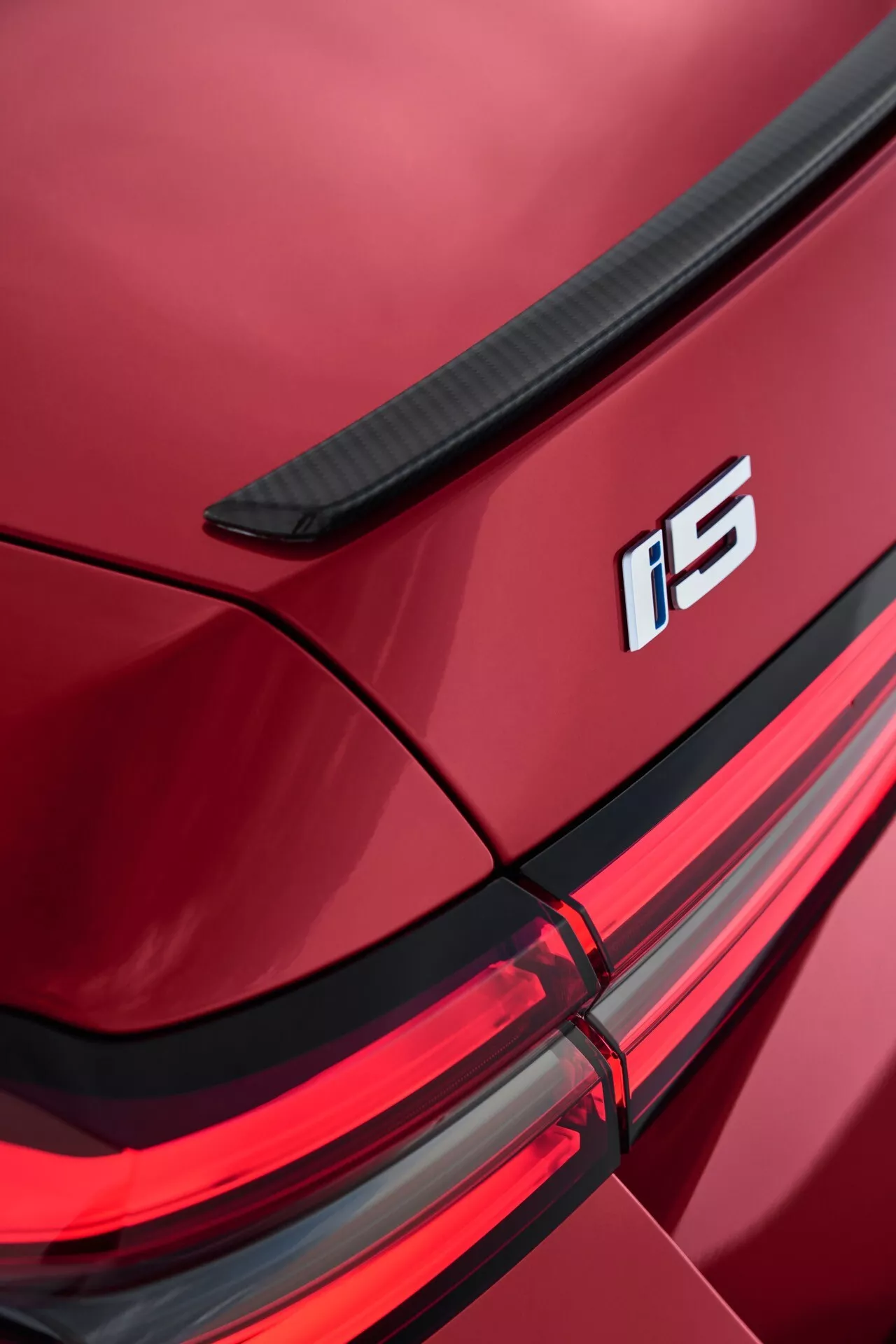 Detailní pohled na pravý zadní roh BMW řady 5 (G60-G61) 2023 s integrovaným spoilerem na víku zavazadlového prostoru, označením i5, sestavou LED zadních světel a lesklým metalickým červeným lakováním