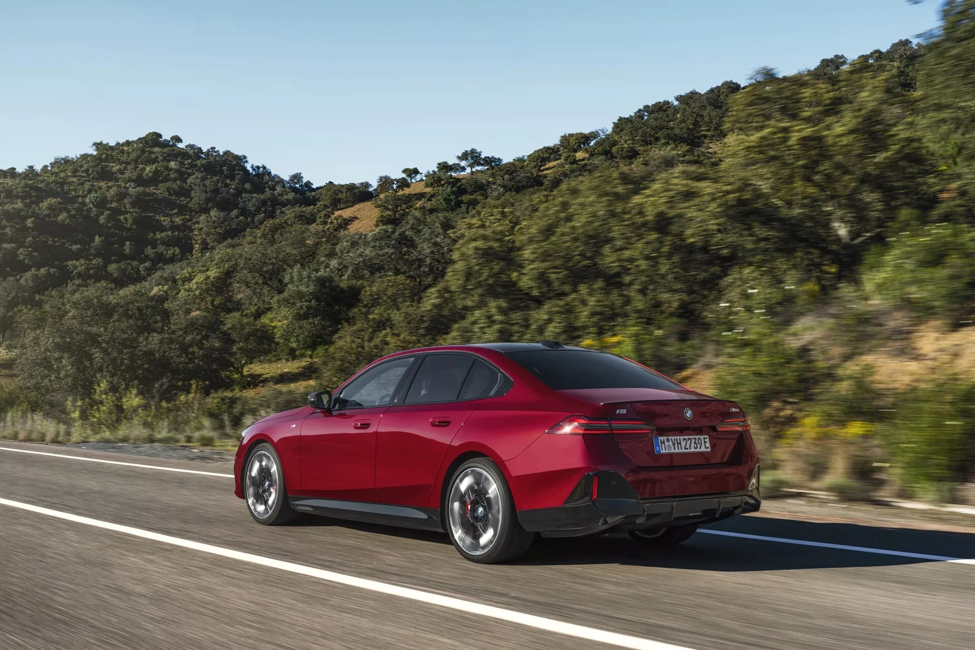 Červený sedan BMW řady 5 (G60-G61) se čtyřmi dveřmi v pohybu zachycený z tříčtvrtečního zadního úhlu, zdůrazňující zadní a boční designové prvky jako tvar kufru, zadní nárazník, LED zadní světla a aerodynamické linie na pozadí horské krajiny pod jasnou oblohou