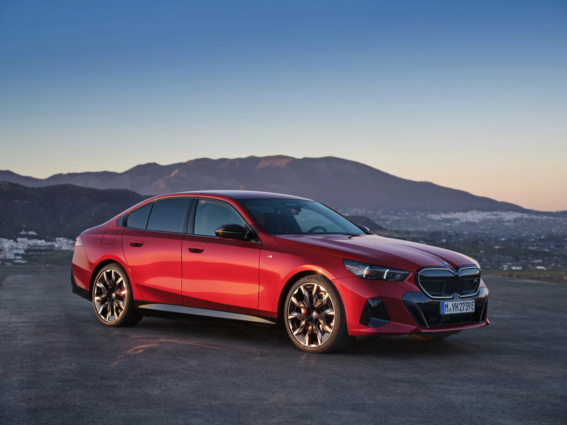 BMW řady 5 z roku 2023 (G60-G61) zobrazené z úhlu tří čtvrtin zepředu, zdůrazňující jeho charakteristickou přední mřížku s logem uprostřed, štíhlé LED světlomety, přední nárazník s integrovanými nasávacími otvory a leštěná kola z lehkých slitin s více paprsky proti dynamickému červenému kovovému finiši a pozadí soumraku