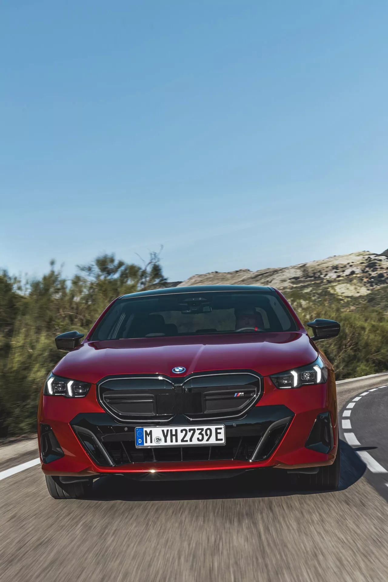 BMW řady 5 (G60-G61) 2023 v červené barvě, předvádějící charakteristickou ledvinovou mřížku, LED světlomety a dynamický přední nárazník z nízkého úhlu zepředu na silnici, což zdůrazňuje rychlost vozidla a pokročilé funkce elektrifikované řady iPerformance