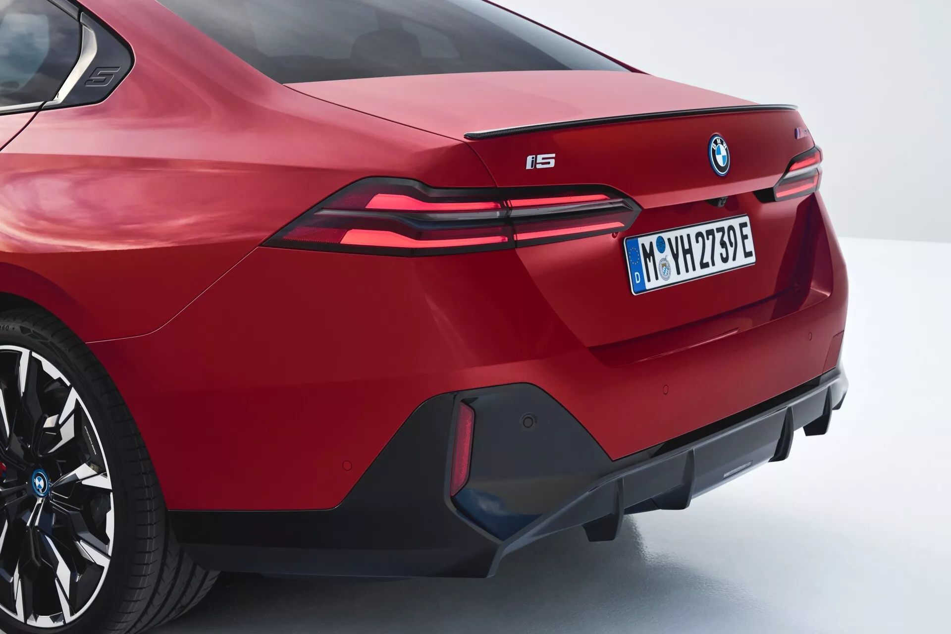 Zadní úhlový pohled na červené BMW i5 2023 ukazující sportovní sedanový design, integrovaný spoiler, výrazné logo BMW, prodloužená LED zadní světla táhnoucí se do zadních blatníků, označení elektrického modelu 'i5', agresivní zadní nárazník s dekorativním difuzorem a falešnými výfuky, ve tříčtvrtečním zadním a bočním profilu.