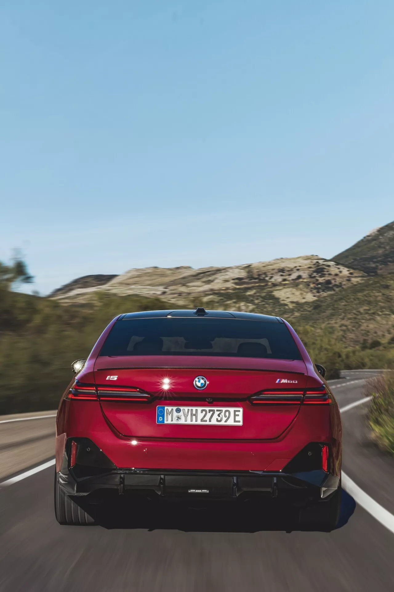 Zadní pohled na jasně červený sedan BMW řady 5 (G60-G61) jedoucí po silnici, s LED zadními světly ve tvaru L, dvojitým výfukovým systémem, odznakem eDrive a parkovacími senzory při jasném denním světle.