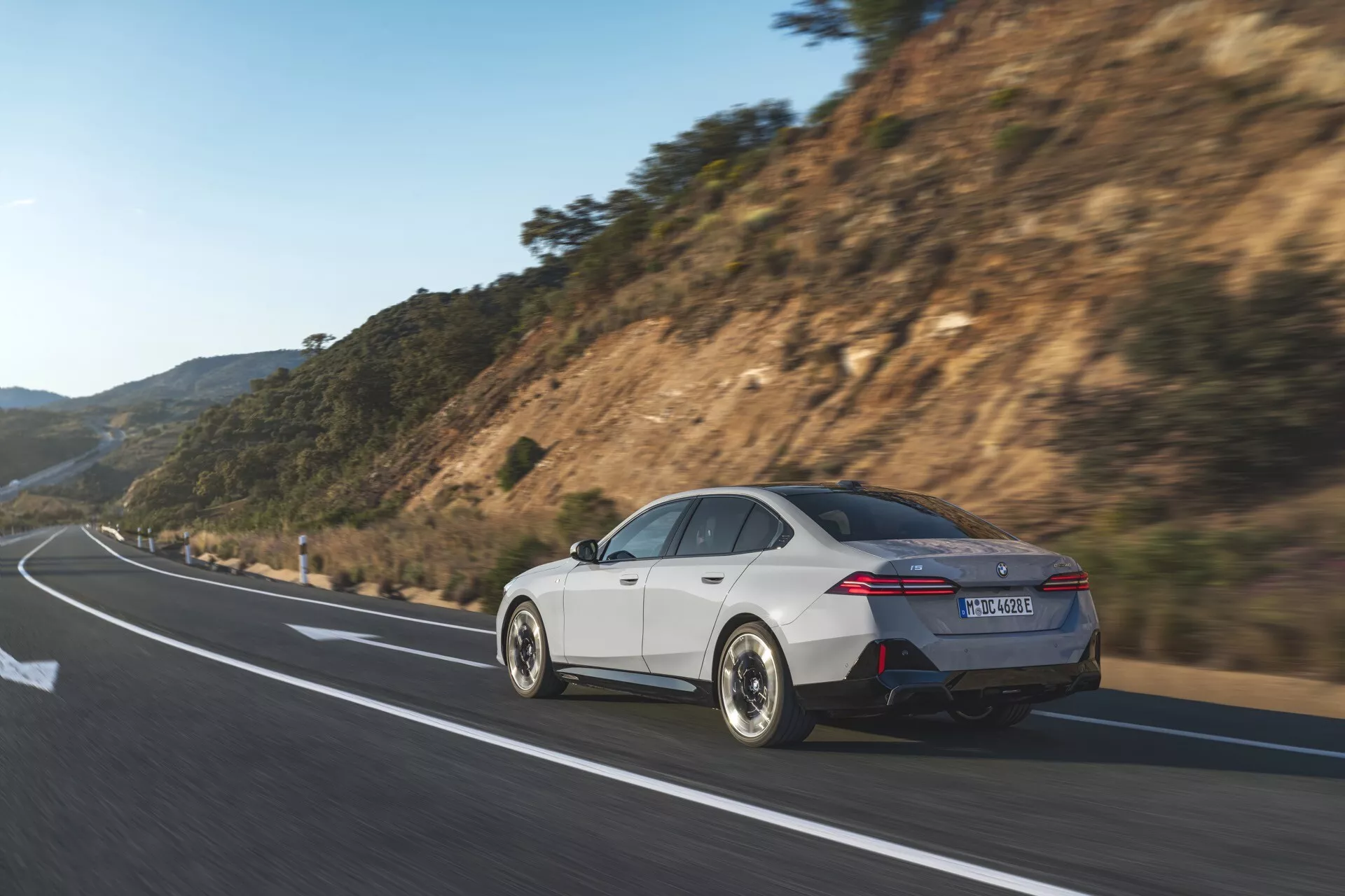 Dynamický pohled zezadu na tři čtvrtiny bílého sedanu BMW řady 5 (G60-G61) z roku 2023, jedoucího po dálnici, který předvádí sportovní design, charakteristická zadní světla, dvojité výfuky, elegantní slitinová kola a aerodynamická boční zrcátka uprostřed přírodního prostředí