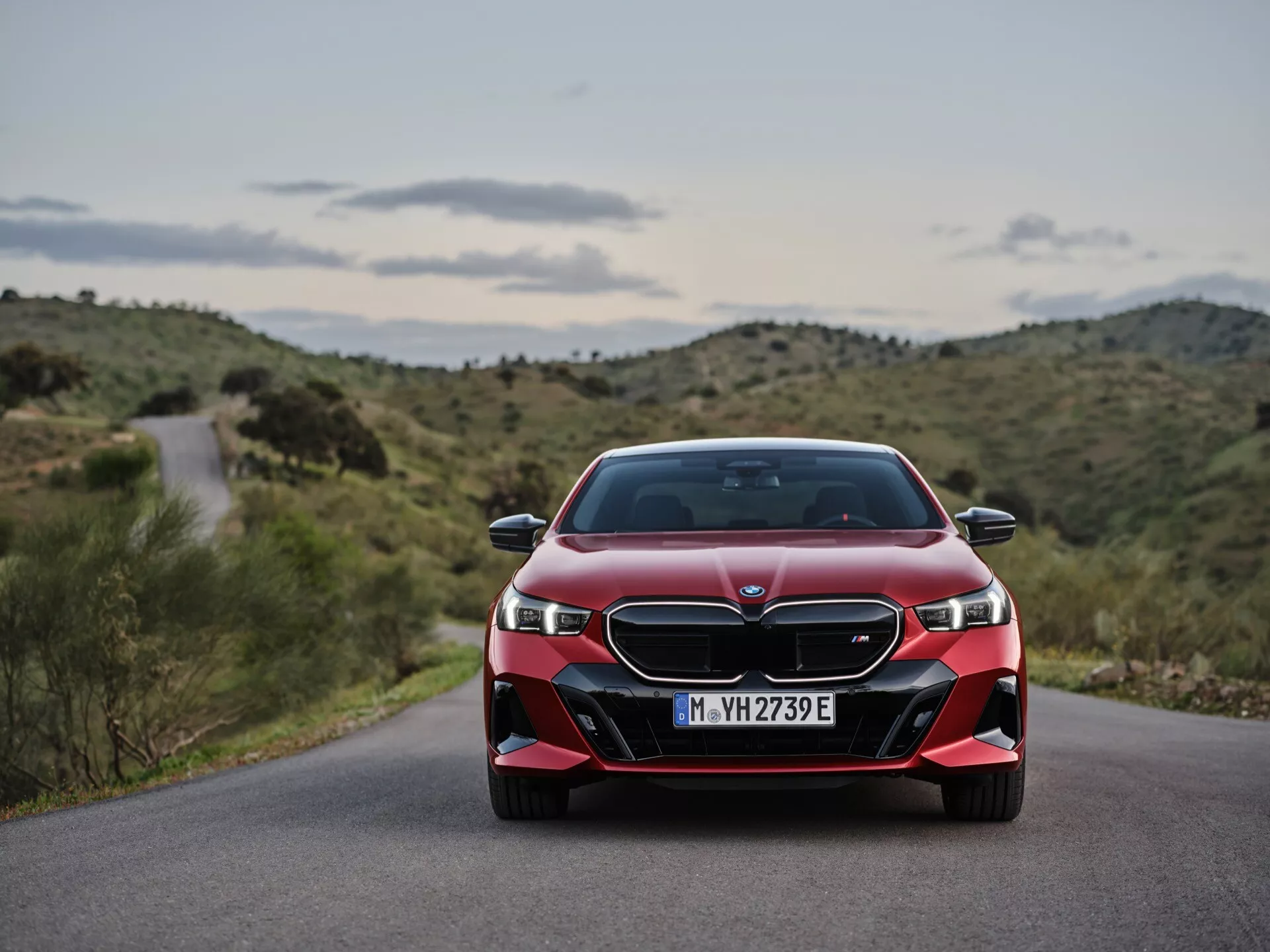 Čelní pohled na červené BMW 5 Series (G60-G61) ročník 2023, zobrazující LED světlomety, dvojitou ledvinkovou mřížku a sportovní nárazník na pozadí klikaté silnice a kopcovité krajiny