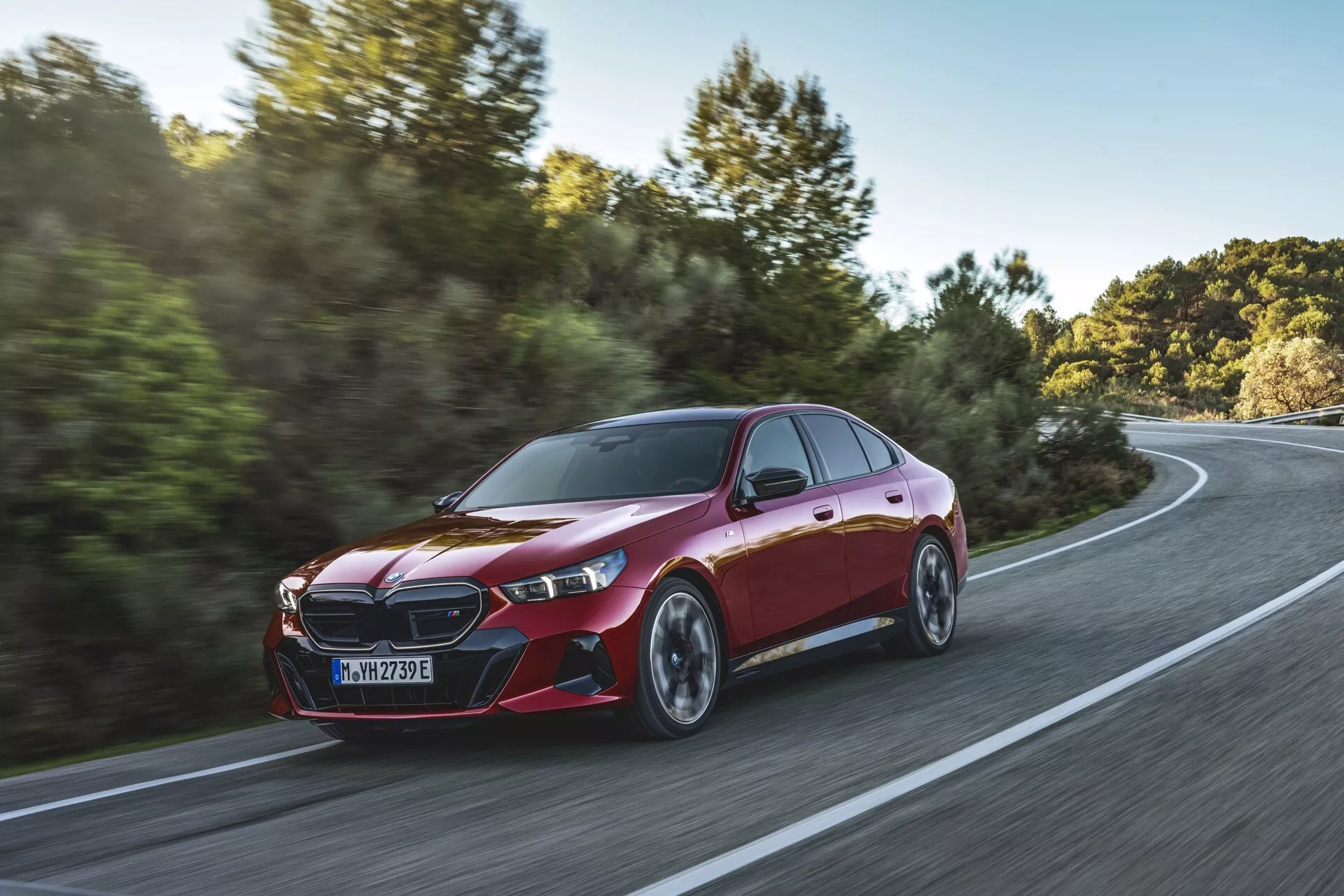 Dynamický tříčtvrteční pohled na BMW řady 5 (G60-G61) z roku 2023 v pohybu na zakřivené cestě uprostřed venkovské krajiny, zdůrazňující jeho metalický červený lak, výraznou přední masku, elegantní LED světlomety, aerodynamická boční zrcátka, sportovní kontury, nízkoprofilové pneumatiky a vícepaprsková slitinová kola