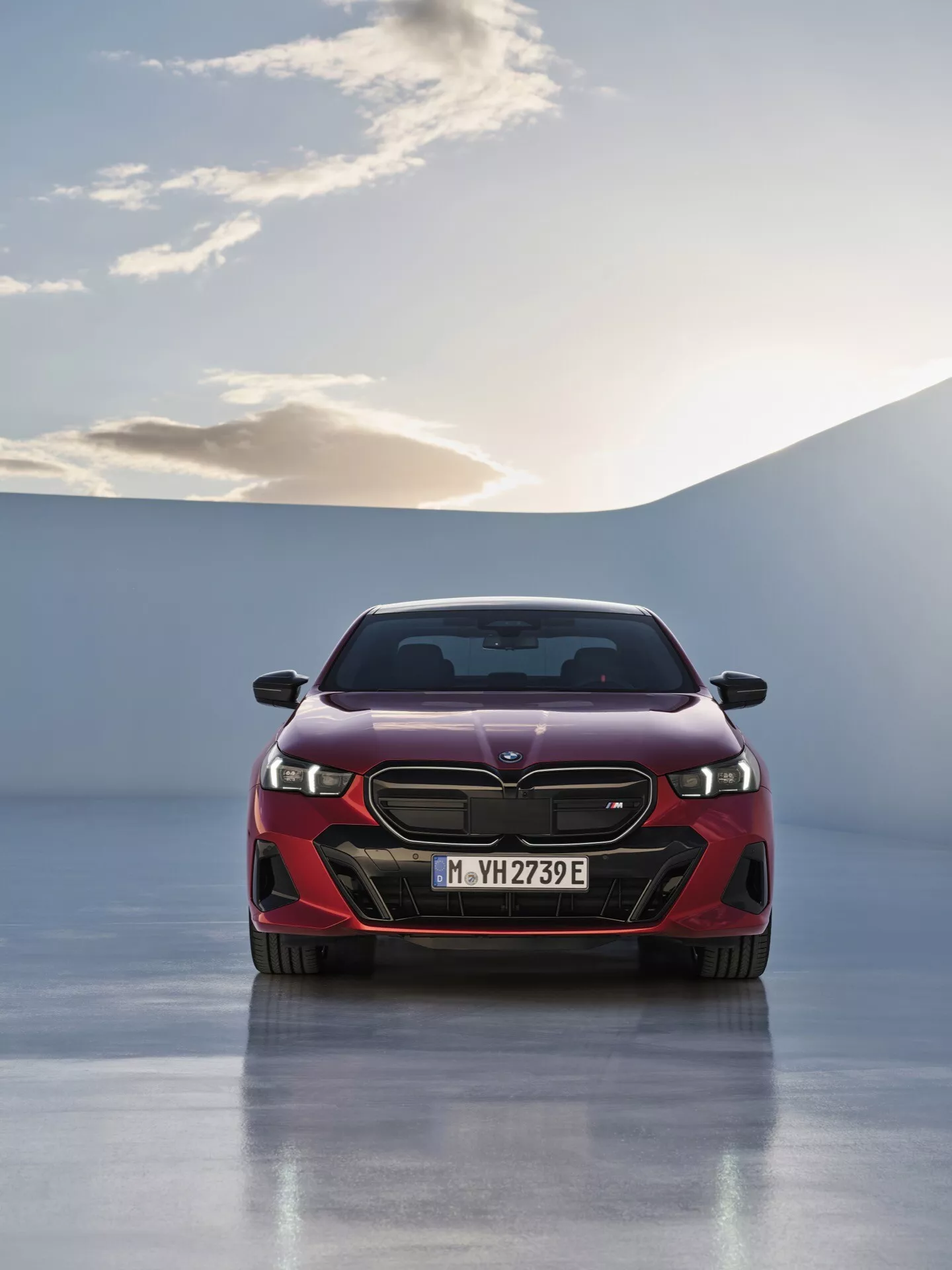 BMW řady 5 (G60-G61) 2023 v nápadné červené barvě předvádějící svůj hladký sportovní design, výraznou mřížku s emblémem, úhlová LED světlomety a aerodynamický přední nárazník na pozadí východu nebo západu slunce s jasnou oblohou