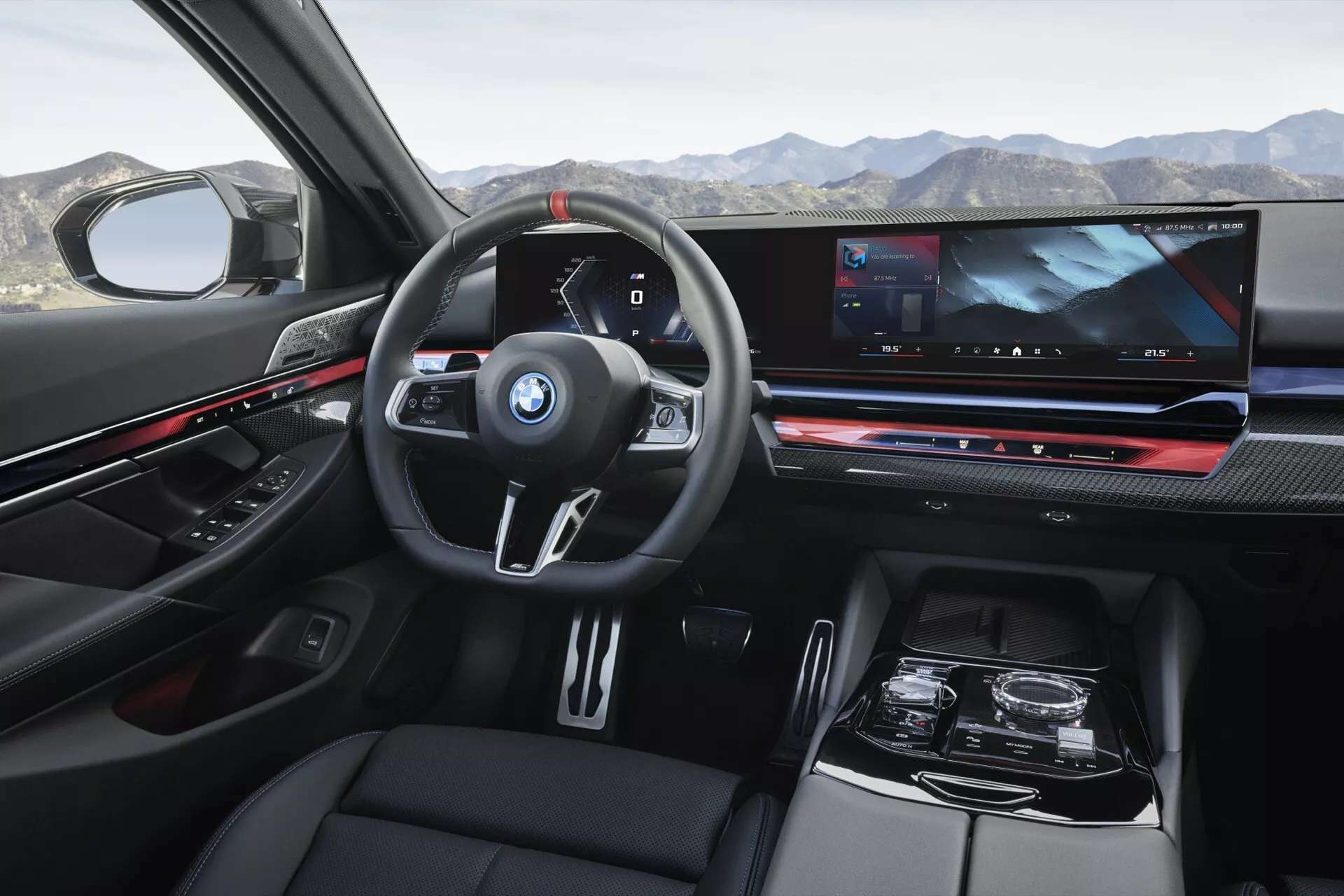 Pohled na interiér BMW řady 5 (G60-G61) z roku 2023 z perspektivy zadního pasažéra ukazující palubní desku, multifunkční volant s logem BMW, digitální přístrojový panel, rozsáhlý informačně-zábavní displej, elektronickou řadicí páku a luxusní povrchy včetně kůže a karbonového dekoru.