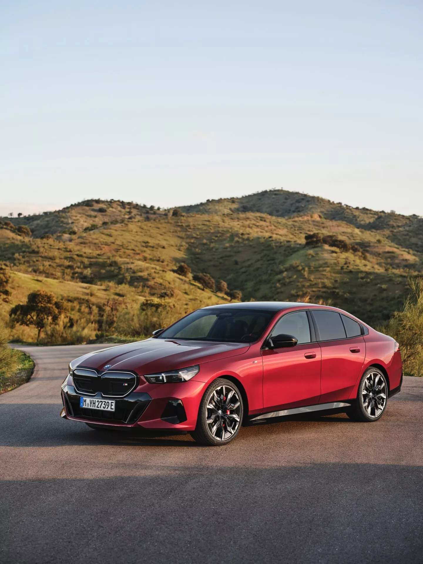 BMW řady 5 2023 (G60-G61) trojčtvrtě čelní pohled na asfaltové cestě s přírodní, kopcovitou krajinou na pozadí, předvádějící LED světlomety, senzory integrované do mřížky, přívody vzduchu, sportovní litá kola s více drážkami, systém bezklíčového vstupu a vysoce kvalitní lesklý povrch