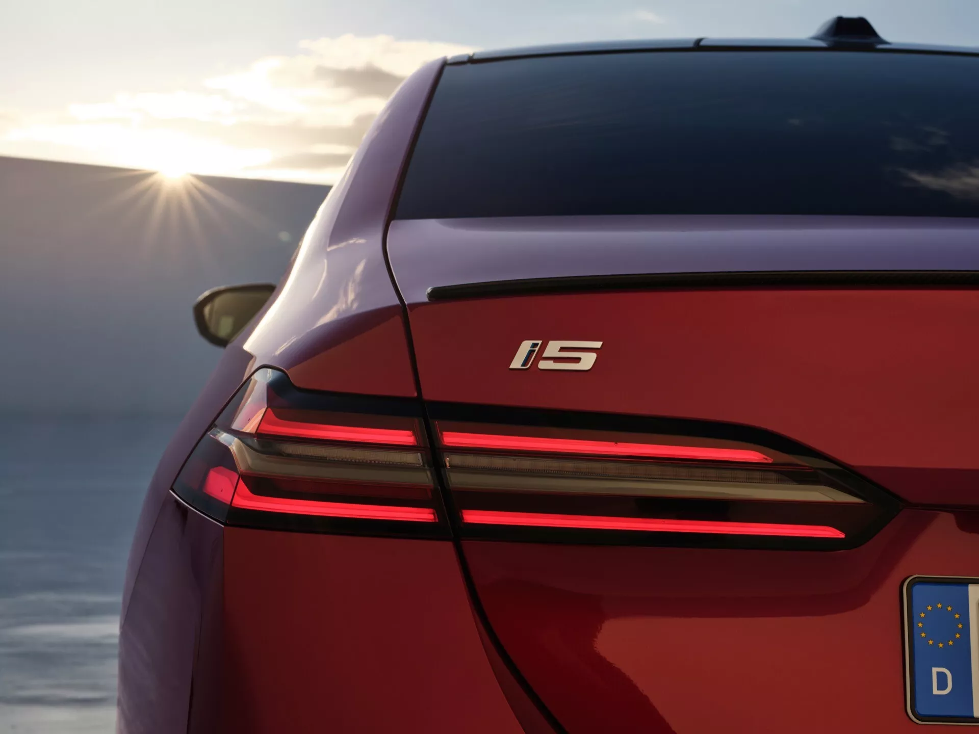 Detailní snímek zobrazující zadní levou část BMW řady 5 (G60-G61) 2023, zdůrazňující její elegantní křivky karoserie, charakteristické celoplošné brzdové světlo, moderní design LED zadních světel a označení 'i5' proti rozmazanému přírodnímu pozadí s teplým slunečním zářením.