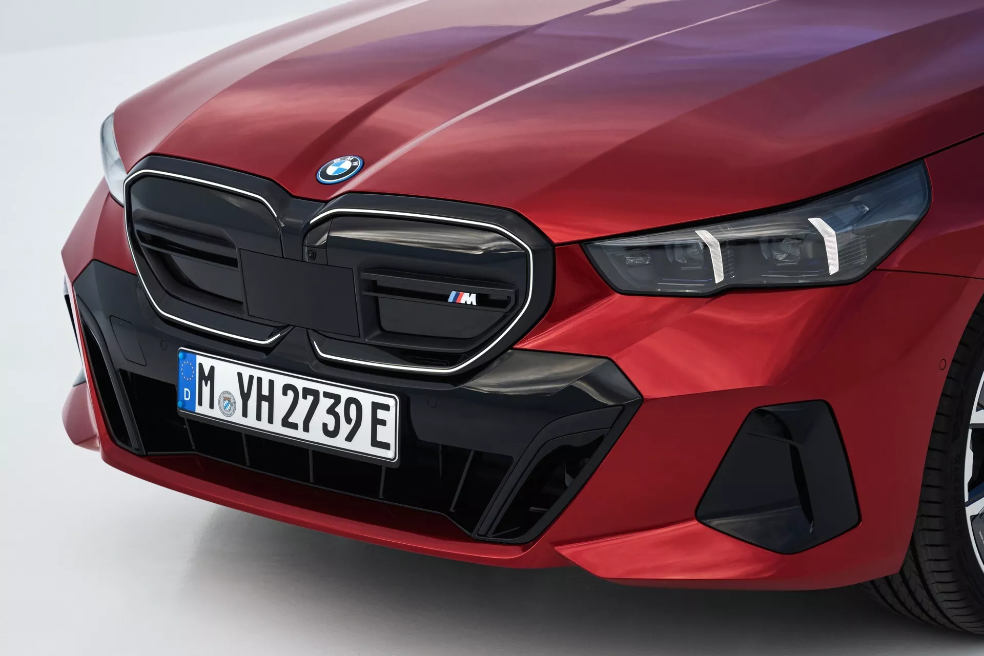 Detailní zkosený pohled na přední levou stranu červeného BMW řady 5 (G60-G61) z roku 2023 s ikonickou ledvinovou mřížkou označenou jako model 'M', světlometovým modulem LED a specializovanými aerodynamickými prvky naznačujícími elektrické nebo hybridní schopnosti