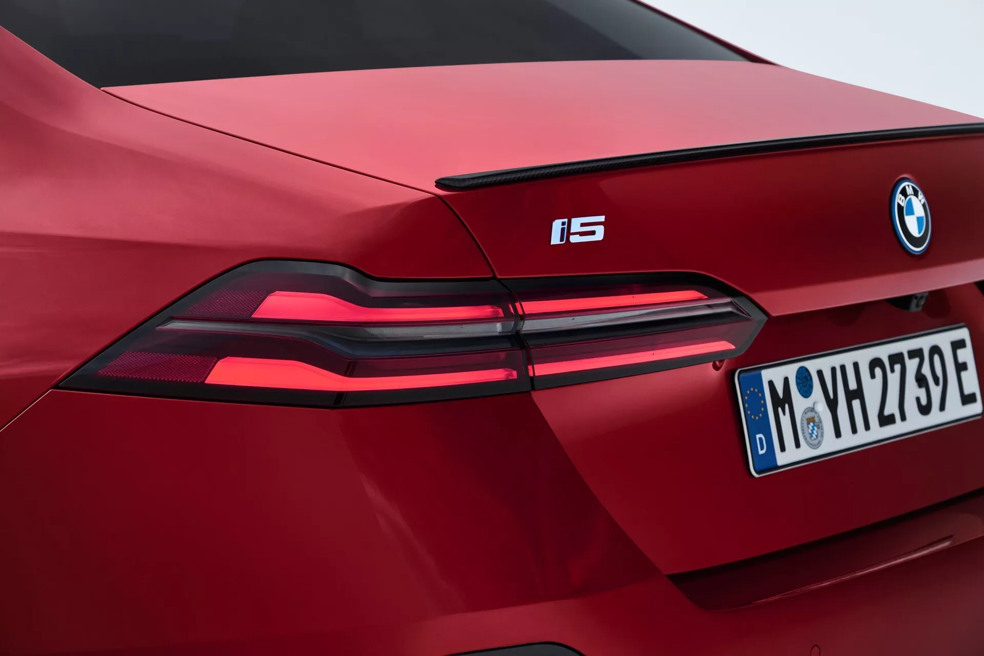 Detailní pohled na část zadní části červeného BMW i5 z roku 2023, zdůrazňující moderní LED zadní světla, výrazné označení BMW a evropskou poznávací značku z Mnichova, s důrazem na vlastnosti elektrického vozidla