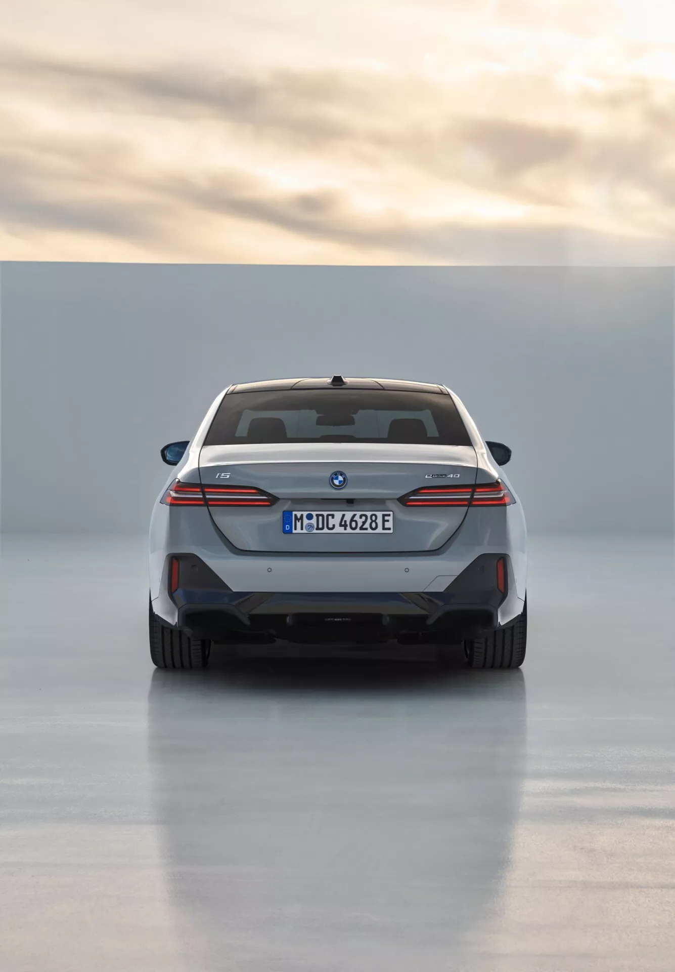 Zadní pohled na BMW řady 5 (G60-G61) rok 2023 předvádějící design celoplošného zadního světla, modré akcenty elektrického modelu, dvojité výfuky a sportovní zadní difuzor, zaregistrované v Mnichově s efektem odrazu na osvětleném podkladu