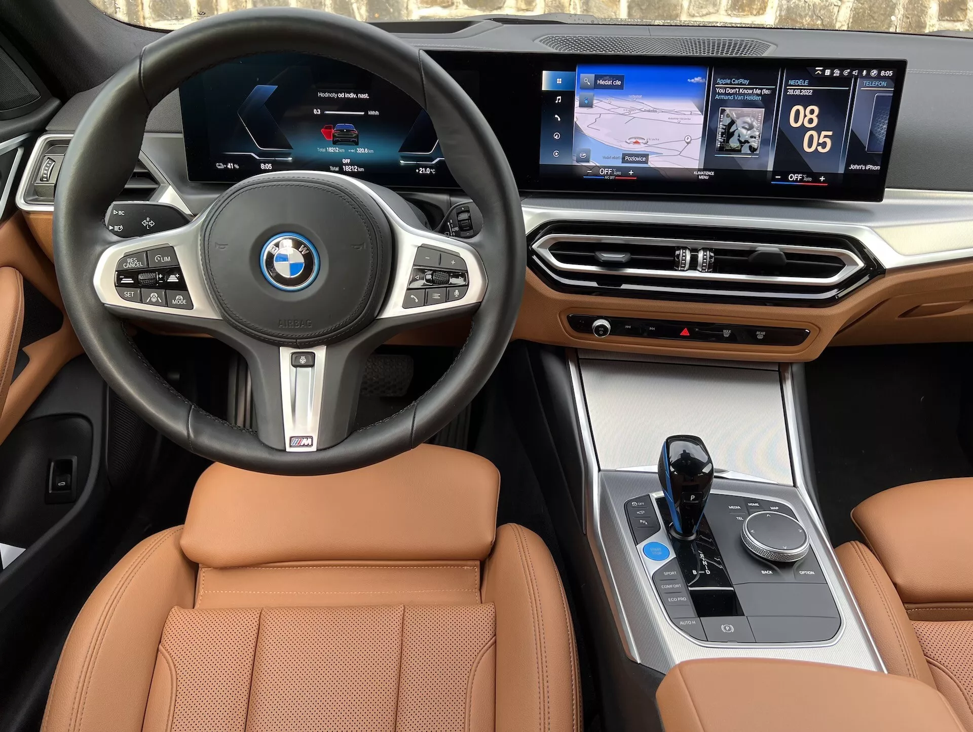 Interiérový pohled na BMW i4 2021, zahrnující centrální volant s logem BMW, multifunkční tlačítka, digitální přístrojový panel, dotykový centrální informační systém zobrazující navigaci a média, luxusní dřevěné akcenty na palubní desce, rozhraní klimatizace, elektronický řadící páčku, ovladač iDrive a sedadla potažená světlou kůží.