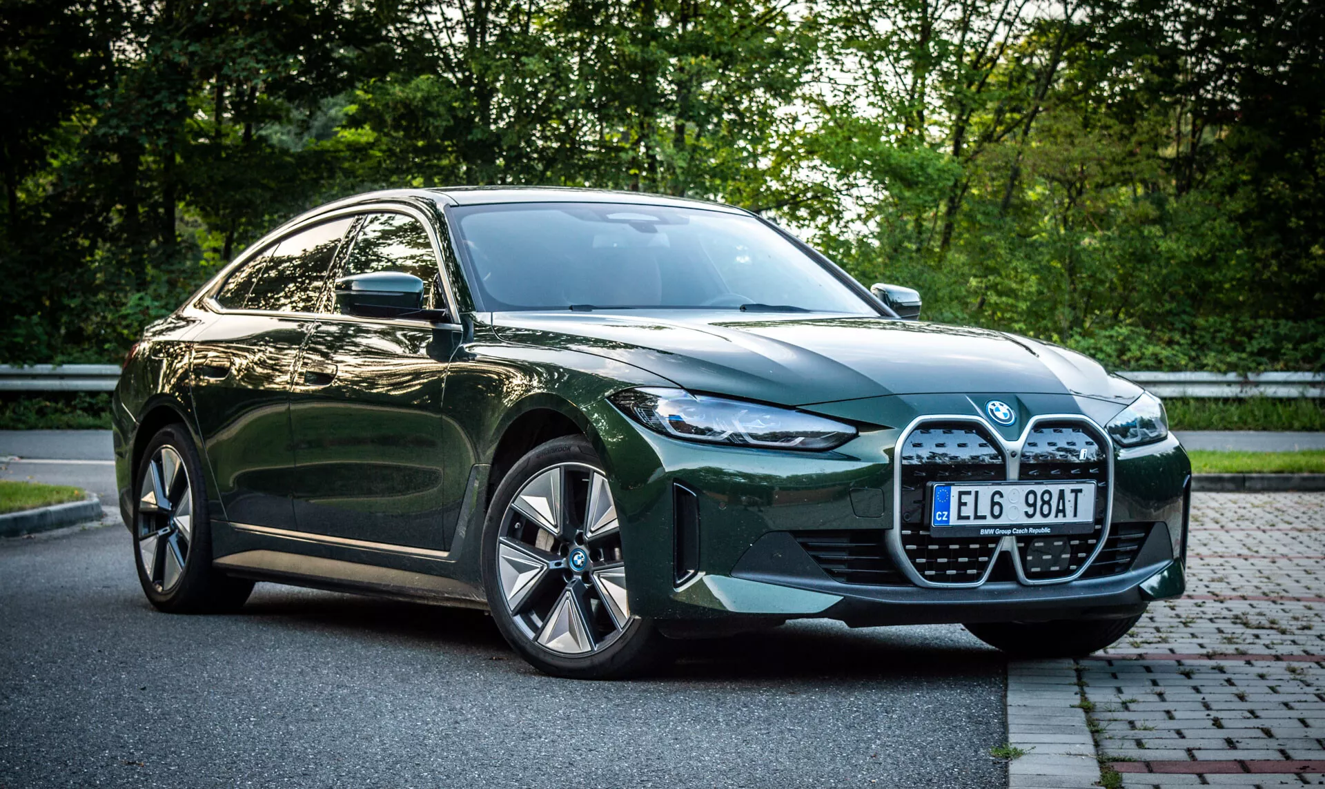 Zelený čtyřdveřový sedan BMW i4 rok 2021 zaparkovaný na cihlové ulici vedle travnaté plochy s listím v pozadí, s výhledem z přední čtvrtiny zobrazujícím velkou masku automobilu, LED světlomety, vstupy vzduchu a slitinová kola