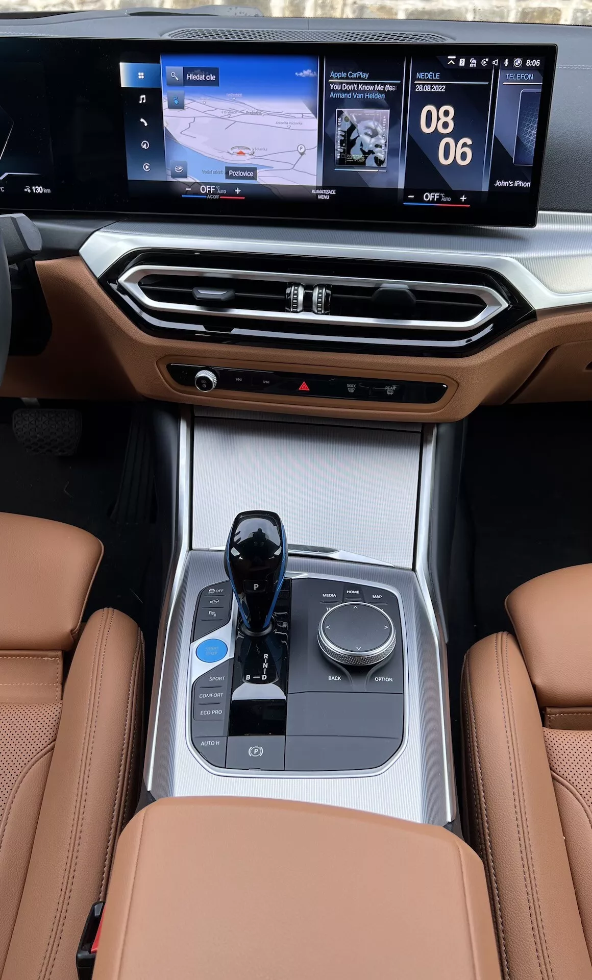 Pohled na středovou konzolu interiéru BMW i4 z roku 2021 ze zadních sedadel, zdůrazňující kožená sedadla, elektronický volič převodovky, ovladač multimédií, držáky nápojů, ovládání klimatizace a dvouúrovňovou infotainment obrazovku s navigací a zobrazením médií