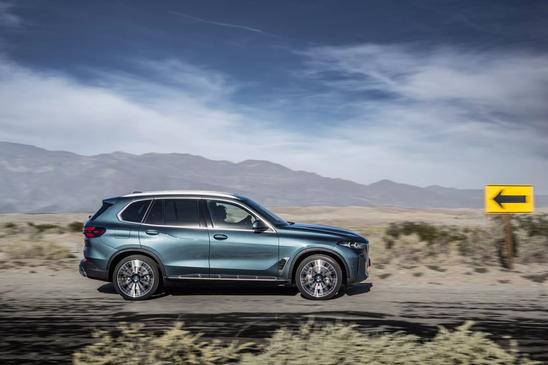 BMW X5 2023 (G05-F95-Facelift) SUV v pohybu, předvádějící aerodynamiku, slitinová kola s více paprsky, integrované kličky dveří, aerodynamická boční zrcátka, tónovaná okna, boční ukazatel, metalický lak a moderní design zadních světel na pozadí rozostřené pouště.