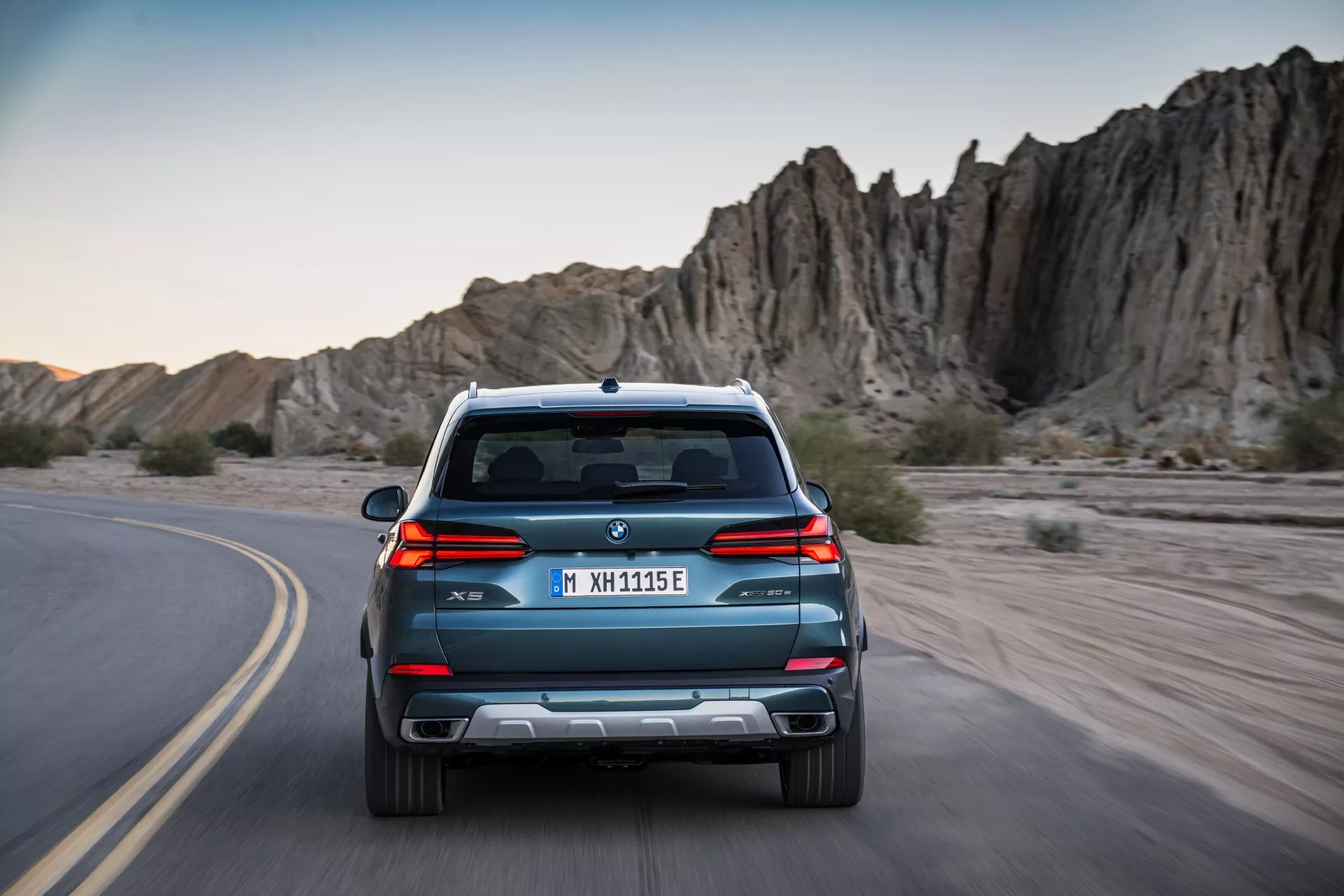 Zadní pohled na BMW X5 G05 F95 facelift 2023, jedoucí po klikaté cestě, s elegantními liniemi, dvojitým výfukem a parkovacími senzory na pozadí dobrodružné skalnaté krajiny