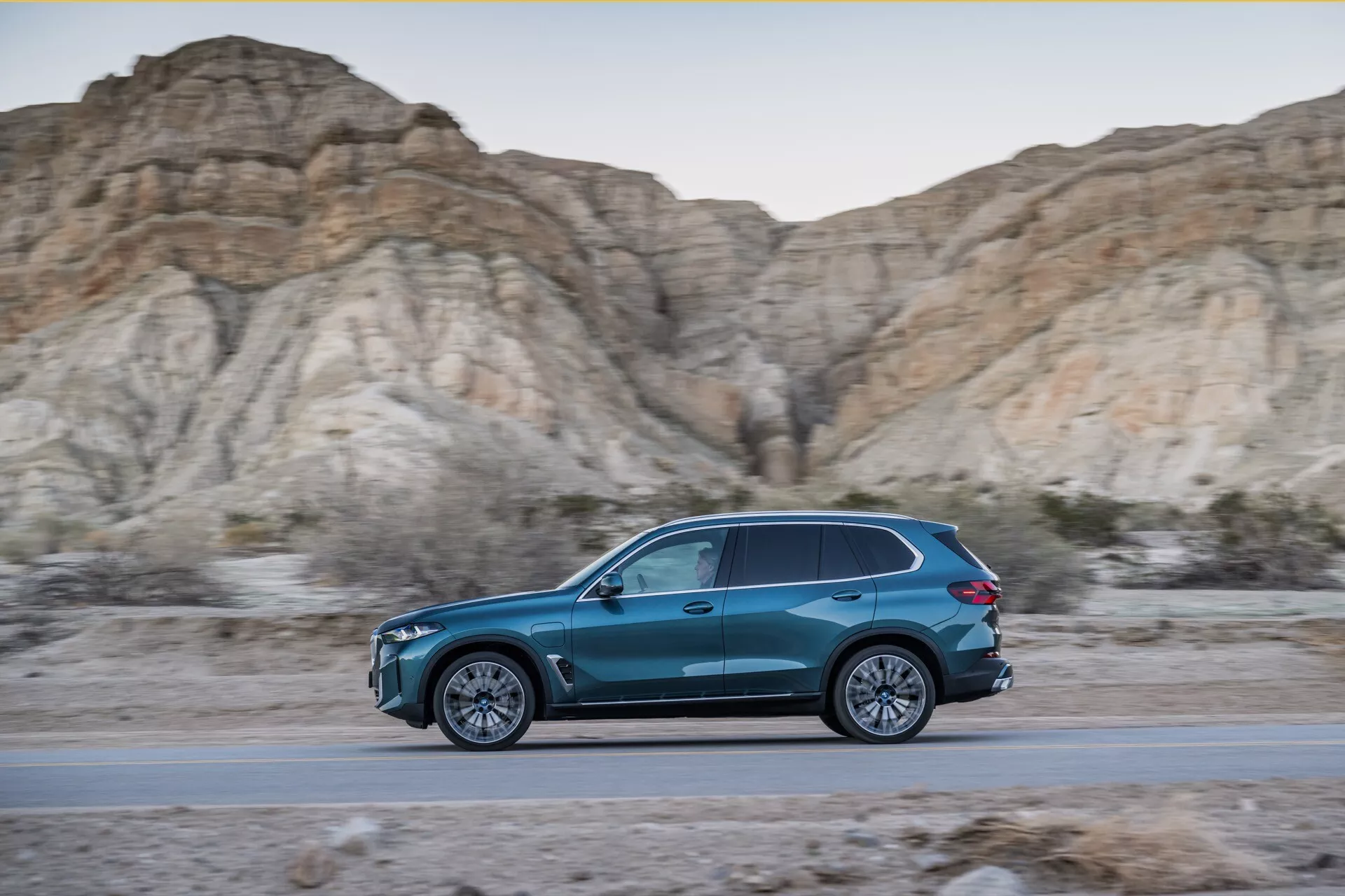 Dynamický boční profil BMW X5 2023 (G05-F95 facelift) v pohybu, modré SUV ve tvaru slzy na silnici s pozadím drsných, nehostinných kopců během zlaté hodiny, předvádějící elegantní design, aerodynamickou účinnost, slitinová kola s více paprsky a LED osvětlení