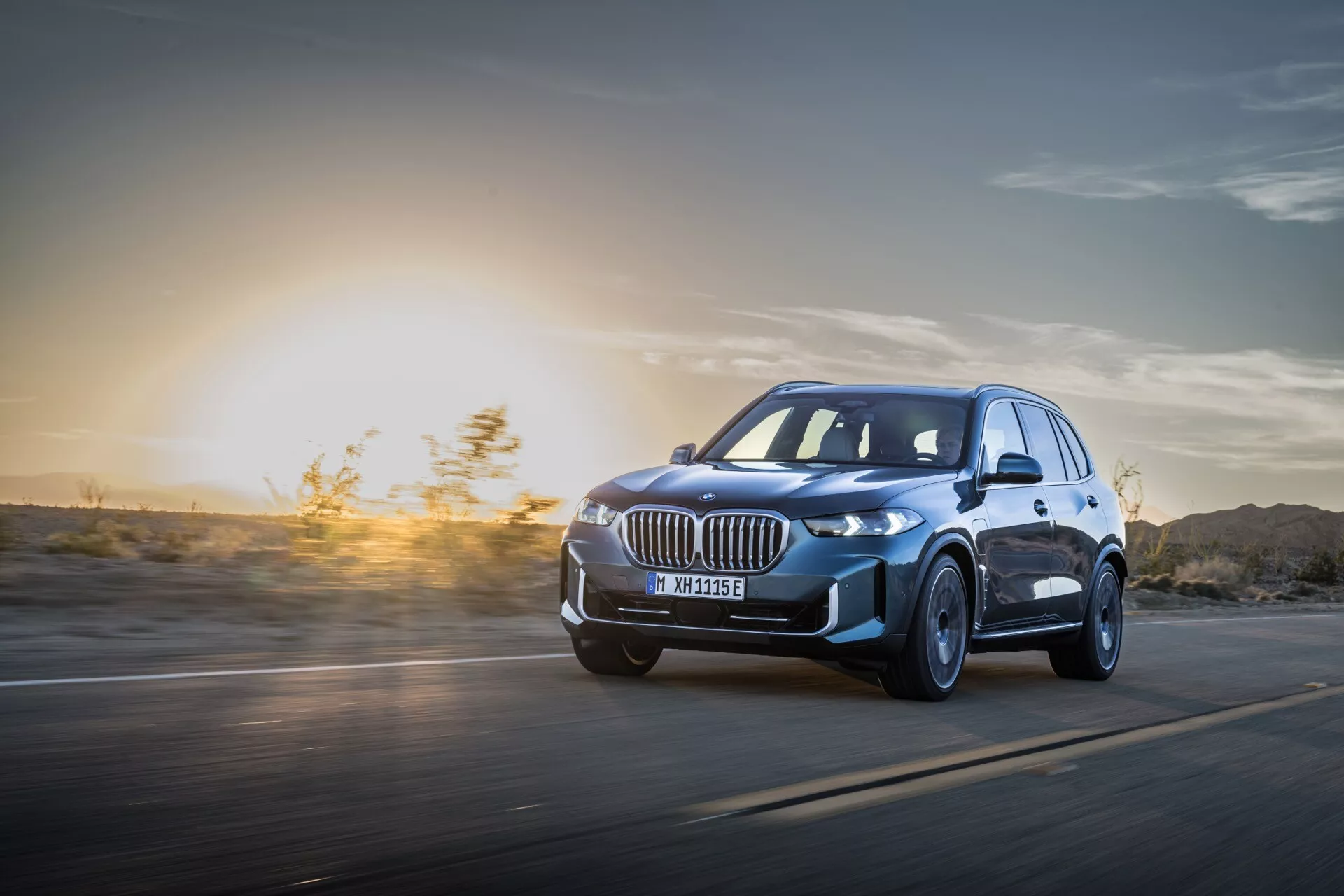 BMW X5 2023 (G05-F95 Facelift) v pohybu: Dynamický pohled z nízkého úhlu zdůrazňující LED světlomety, slitinová kola a aerodynamický design na pozadí západu slunce