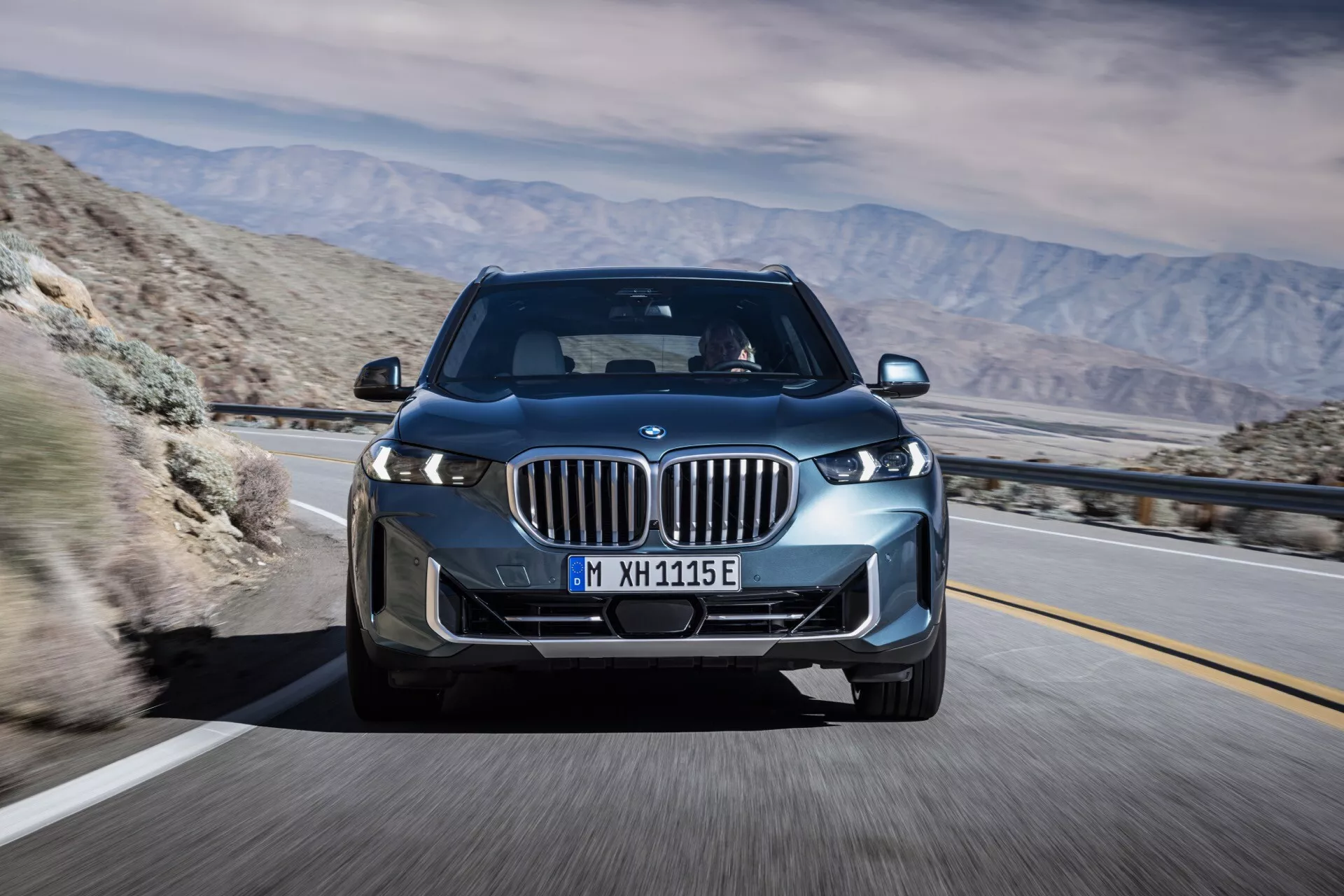 BMW X5 2023 (G05 F95 facelift) v pohybu, zachycené z předního tříčtvrtečního úhlu, zvýrazňující jeho LED světlomety, ledvinkovou mřížku s aktivními žaluziemi, slitinová kola a elegantní aerodynamický design na pozadí horské pouště, naznačující pokročilou technologii a potenciální elektrické nebo hybridní schopnosti