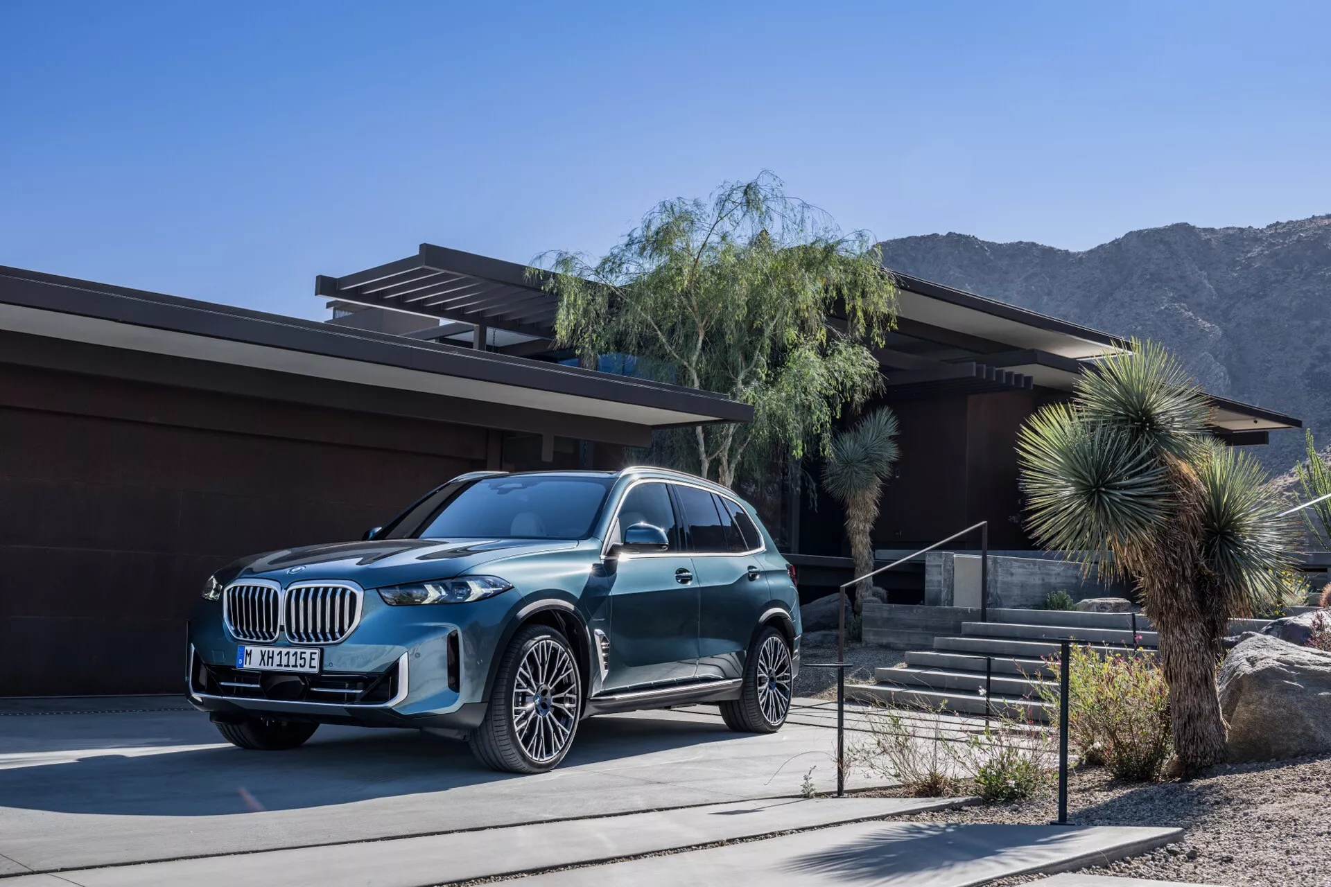 Luxusní SUV BMW X5 2023 (G05-F95 Facelift) zaparkované před moderním domem se zázemím pouštní a horské krajiny, s metalickým povrchem, velkou mřížkou, LED světlomety a litými koly s vícepaprskovým designem