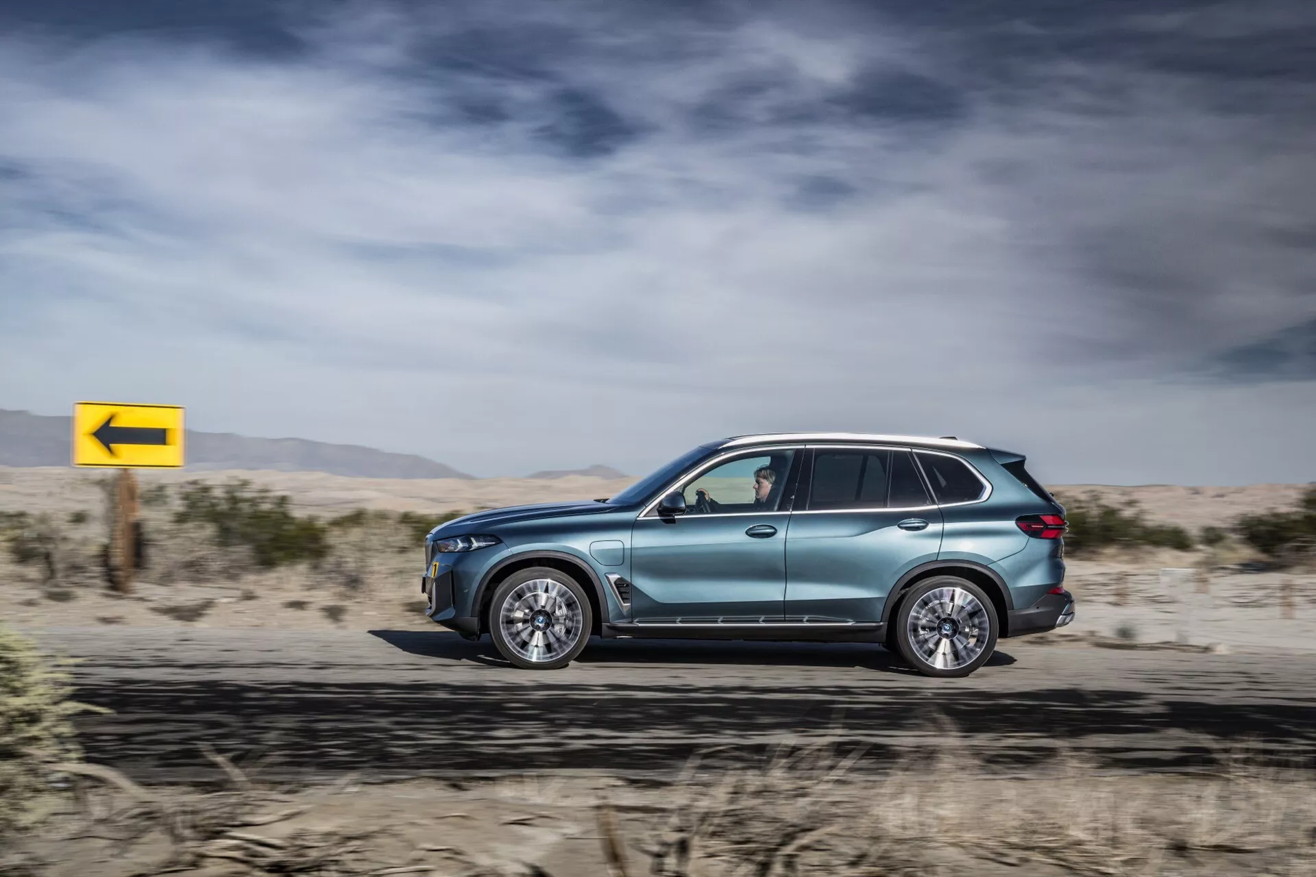 Boční profil BMW X5 2023 (G05-F95 Facelift) v pohybu, prezentující metalický lak, aerodynamický design, luxusní slitinová kola, tónovaná okna se zrcátky s ukazateli směru, střešní lišty a pokročilou LED technologii, na pozadí pouště s dynamickou žlutou značkou pro levotočení.