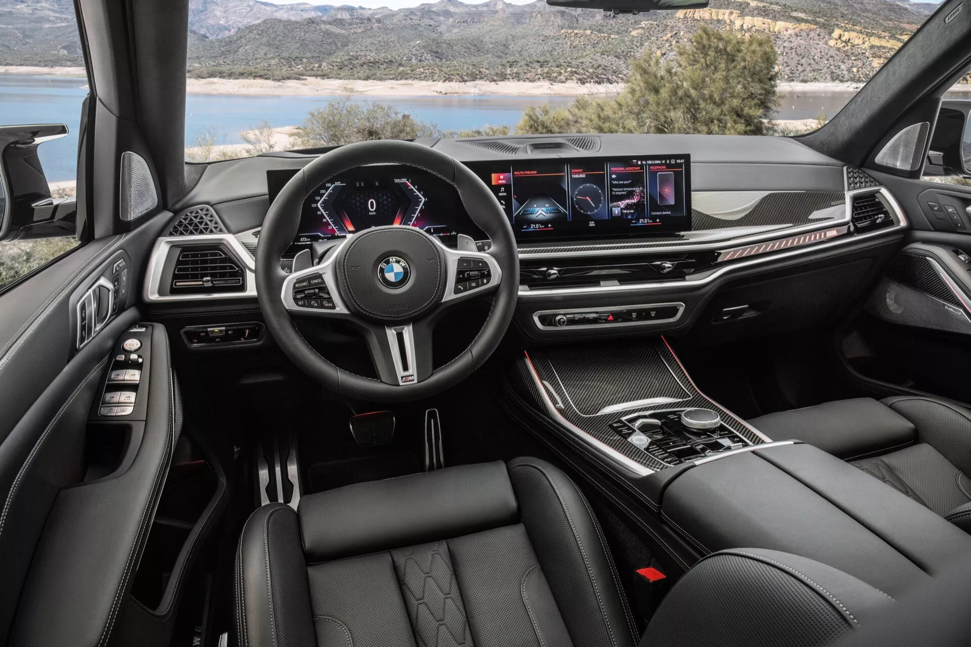 Interiérový pohled na BMW X7 2022 (G07 Facelift) z perspektivy řidiče s multifunkčním volantem s logem BMW, digitálním přístrojovým panelem, širokoúhlým displejem infotainmentu, tlačítky klimatizace, selektorem automatické převodovky, luxusními materiály, sportovně pohodlnými sedadly a ambientním osvětlením