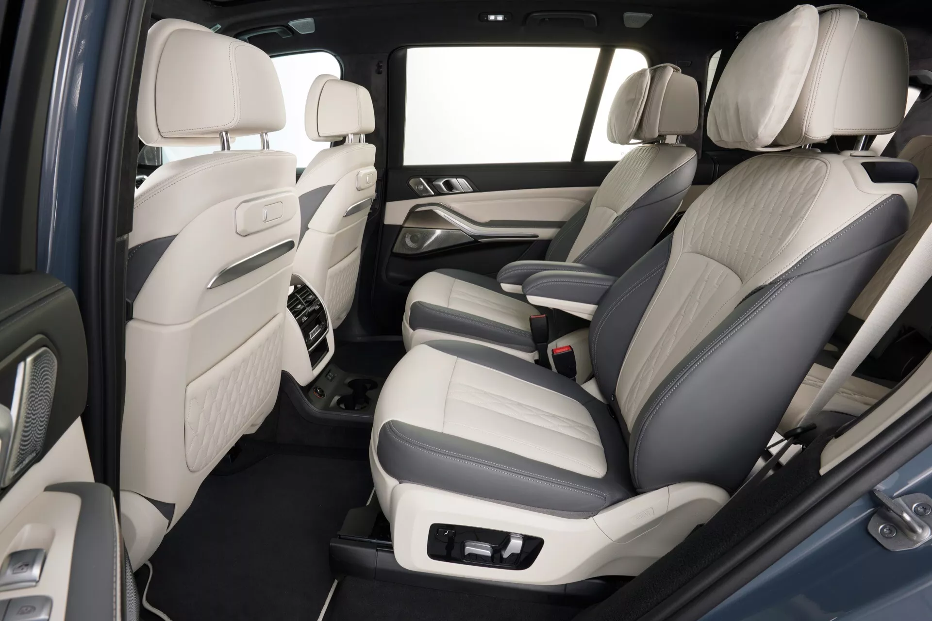 Interiér BMW X7 (G07 Facelift) pro rok 2022 s výhledem na zadní cestující oddíl, s čalouněním z dvoubarevné kůže, pokročilými ovládacími prvky audio systému na panelech dveří, středovou konzolou s akcenty z kvalitních materiálů, kobercovou podlahou, prostornou nožní částí a světlým schématem interiéru pro pocit prostorné kabiny