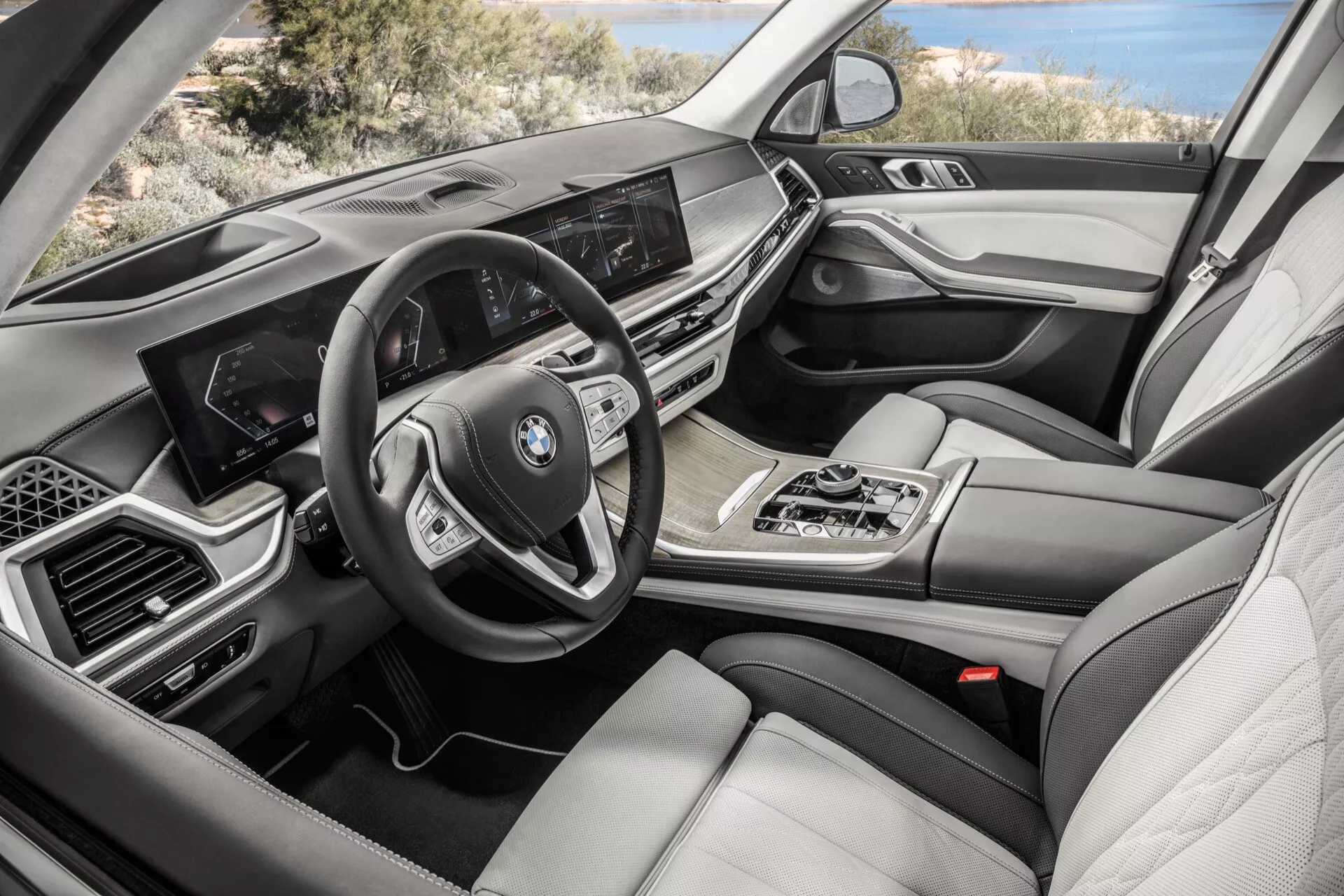 Pohled na interiér BMW X7 z roku 2022 (G07-Facelift) ze strany spolujezdce, zobrazující palubní desku s obrazovkou infotainmentu, multifunkční volant, automatickou převodovku, ovládání multimédií iDrive, dvouzónovou klimatizaci, luxusní kožená sedadla, dřevěné a kovové akcenty a digitální přístrojový panel.