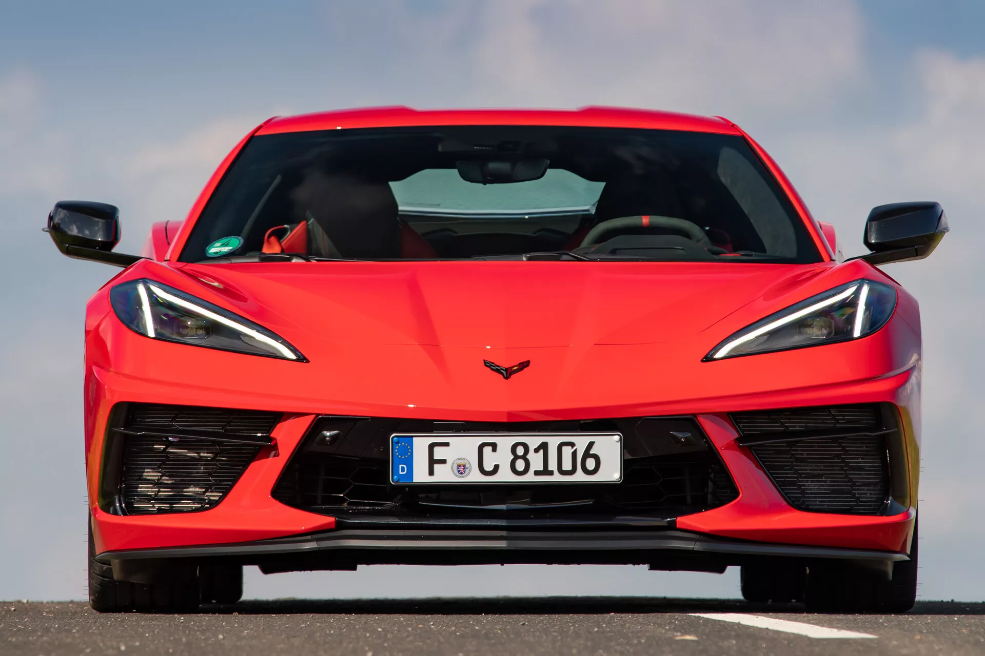 Přední pohled na červený sportovní vůz Chevrolet Corvette 2021 (C8) pořízený z úrovně očí, ukazuje symetrický design vozu. Obrázek se zaměřuje na podrobné čelní prvky vozidla včetně ostrých, úhlových LED světlometů, jedinečné mřížky s malými požadavky na chlazení a velkých vstupních větráků na dolním nárazníku. Emblém Corvette je viditelný na kapotě. Tento detailní snímek zachycuje moderní design a pokročilé technologické prvky vozu.