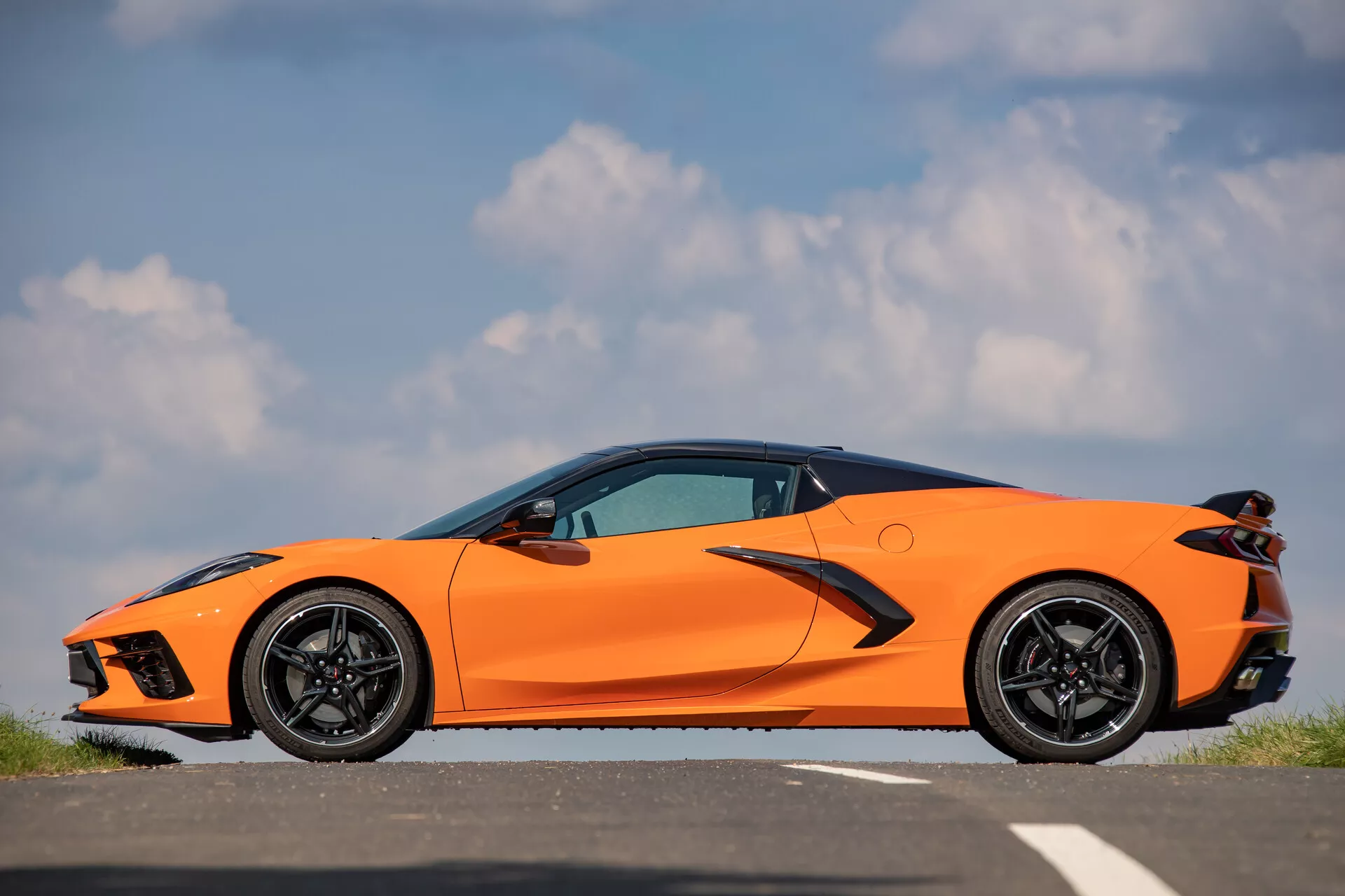 Boční profil Chevroletu Corvette (C8) z roku 2021 v jasně oranžové barvě na asfaltovém povrchu, zobrazující jeho aerodynamicky tvarovanou přední část s ostrými LED světly, velkými přívody vzduchu, elegantní siluetu s výraznou charakterovou linií, zapuštěnými klikami dveří, účelně navrženými bočními prahy, velkými černými sportovními koly s nízkoprofilovými výkonnými pneumatikami, vysoce výkonnými brzdovými třmeny a částečně viditelnými ostrými zadními světly proti jasné obloze.