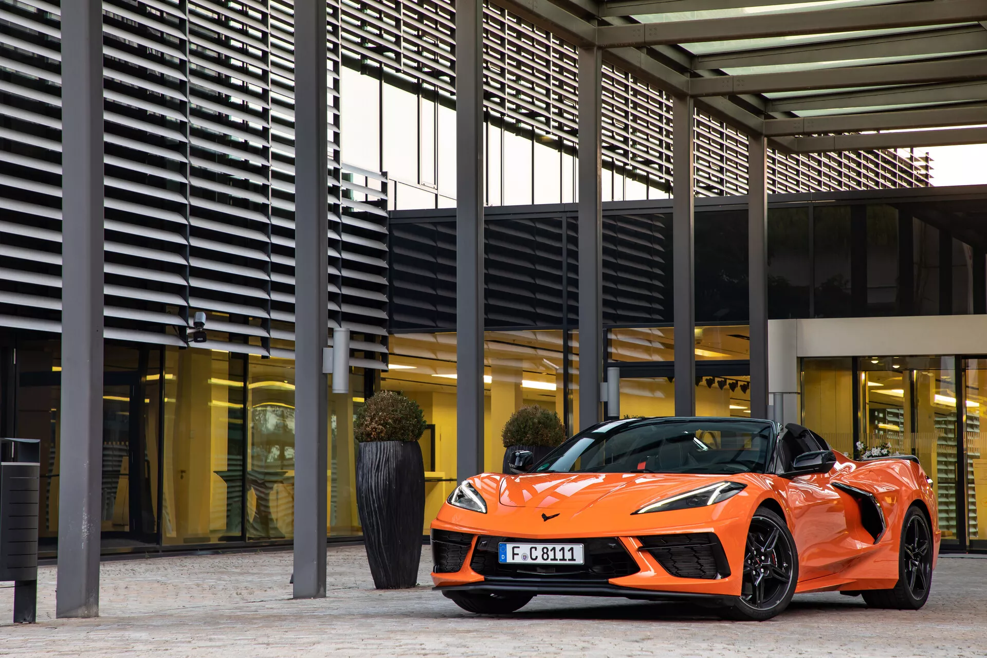 Sportovní oranžový kabriolet Chevrolet Corvette 2021 (C8) zaparkovaný u moderního komplexu budov s výraznými obdélníkovými vzory trámů a sloupů; tříčtvrteční pohled zepředu zvýrazňující stranu řidiče, elegantní světlomety, LED denní světla, nízkoprofilové pneumatiky na vícepaprskových litých kolech a aerodynamické designové prvky. Reflexní povrchy odrážejí lesklý lak a architektonickou integraci.