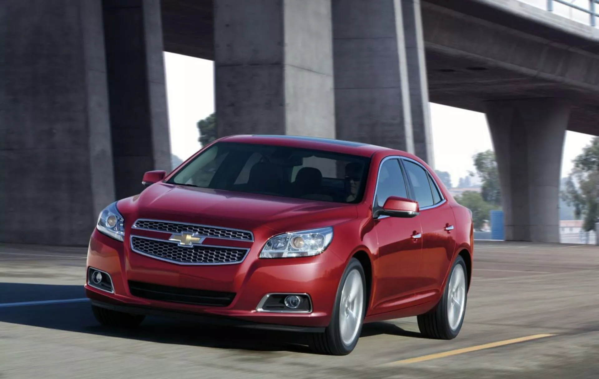 Dynamický pohled na červený sedan Chevrolet Malibu 2012 v pohybu na mostě, zobrazující přední design s chromovanou mřížkou, štíhlými světlomety, sportovní siluetou a slitinovými koly.