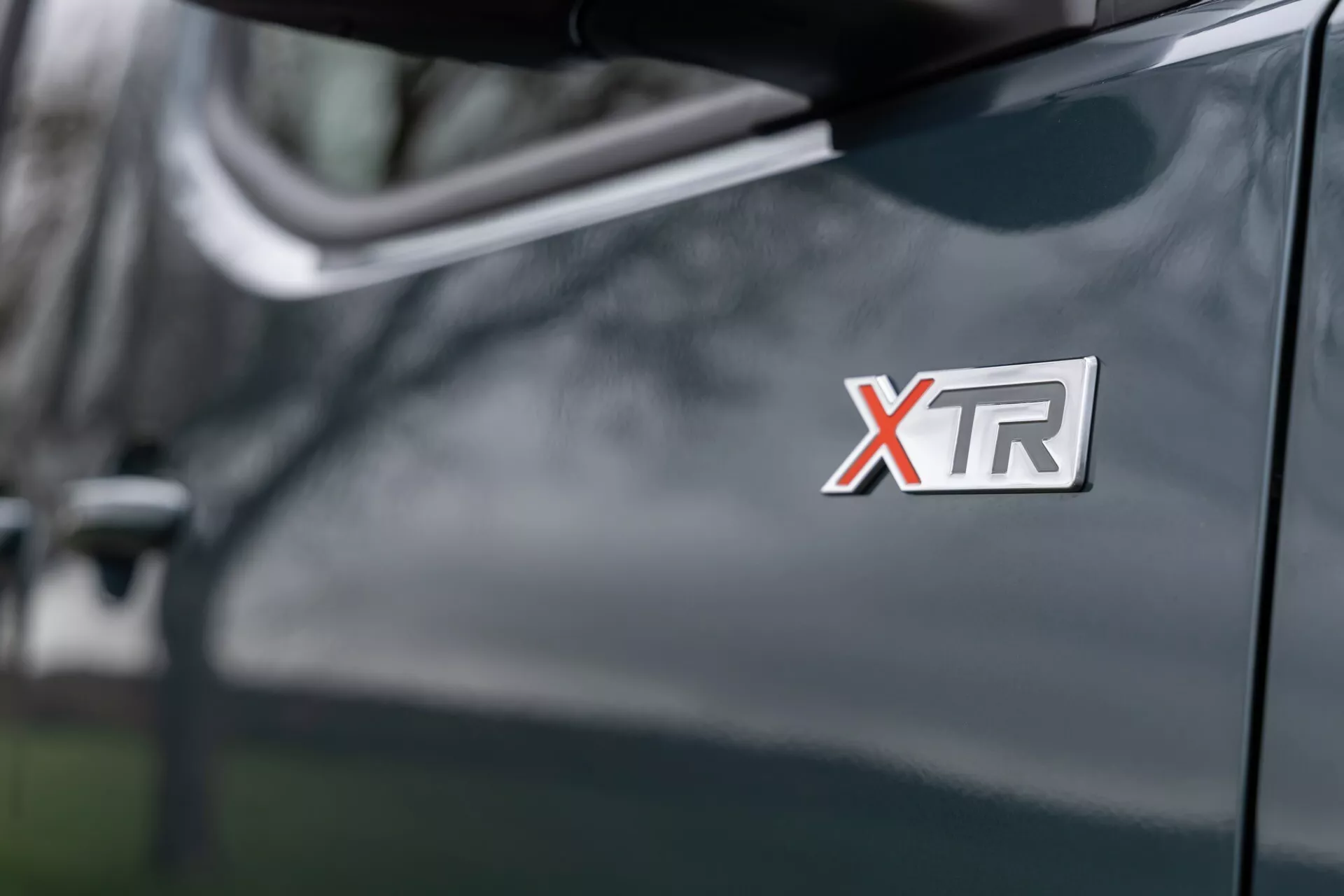 Detailní záběr na odznak XTR Citroen Berlingo 2024 na předním blatníku, předvádějící lesklý tmavě odstínovaný povrch s bočním zrcátkem a rozostřenými odrazy v pozadí.