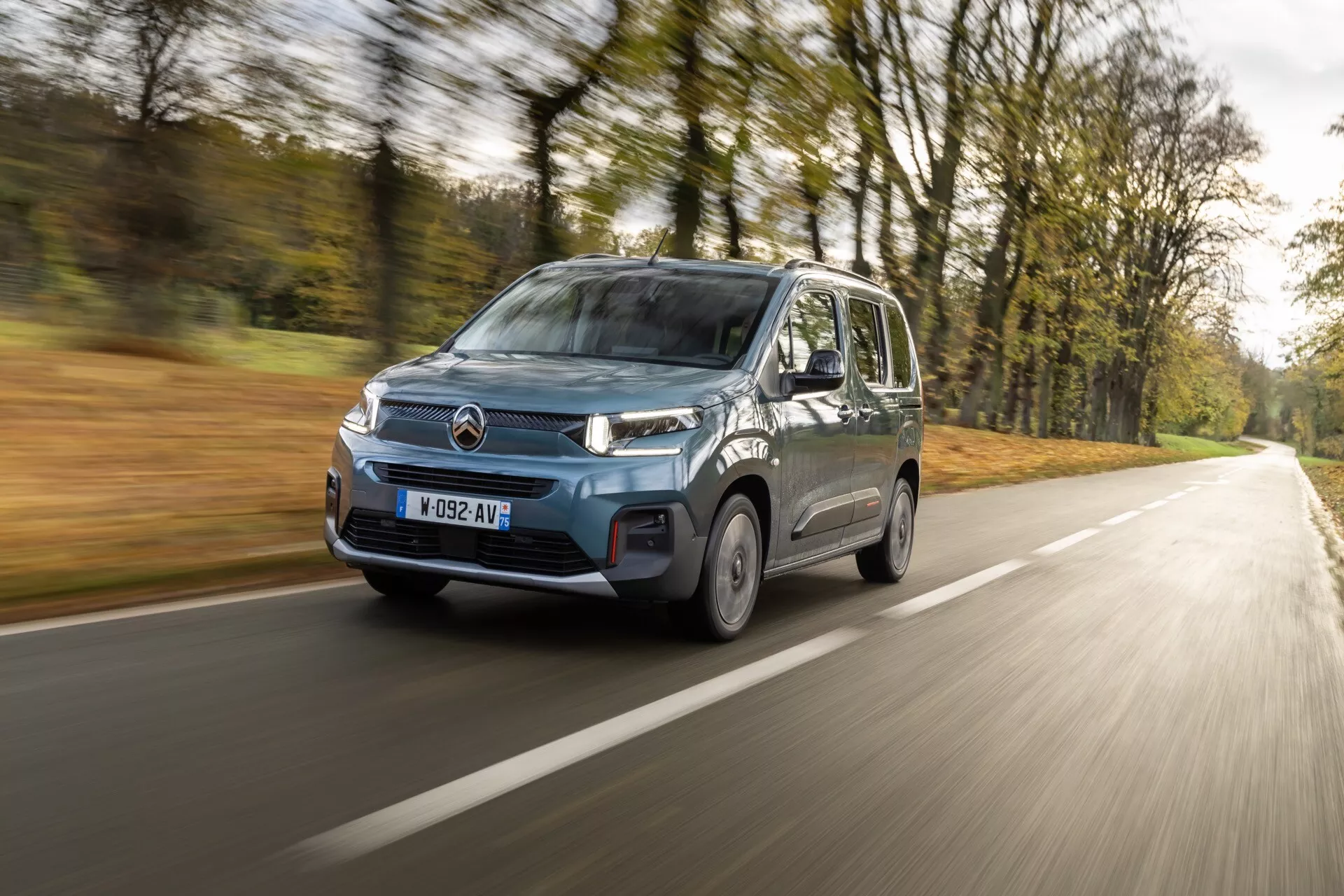 Model Citroen Berlingo 3. generace s faceliftem z roku 2024 v pohybu na venkovské silnici, předvádí moderní design, LED světlomety, mlhová světla a charakteristickou mřížku chladiče, snímek z nízkého až středního úhlu zdůrazňující přední levý kvadrant a boční profil vozidla na pozadí s jemným světlem a alejí stromů.